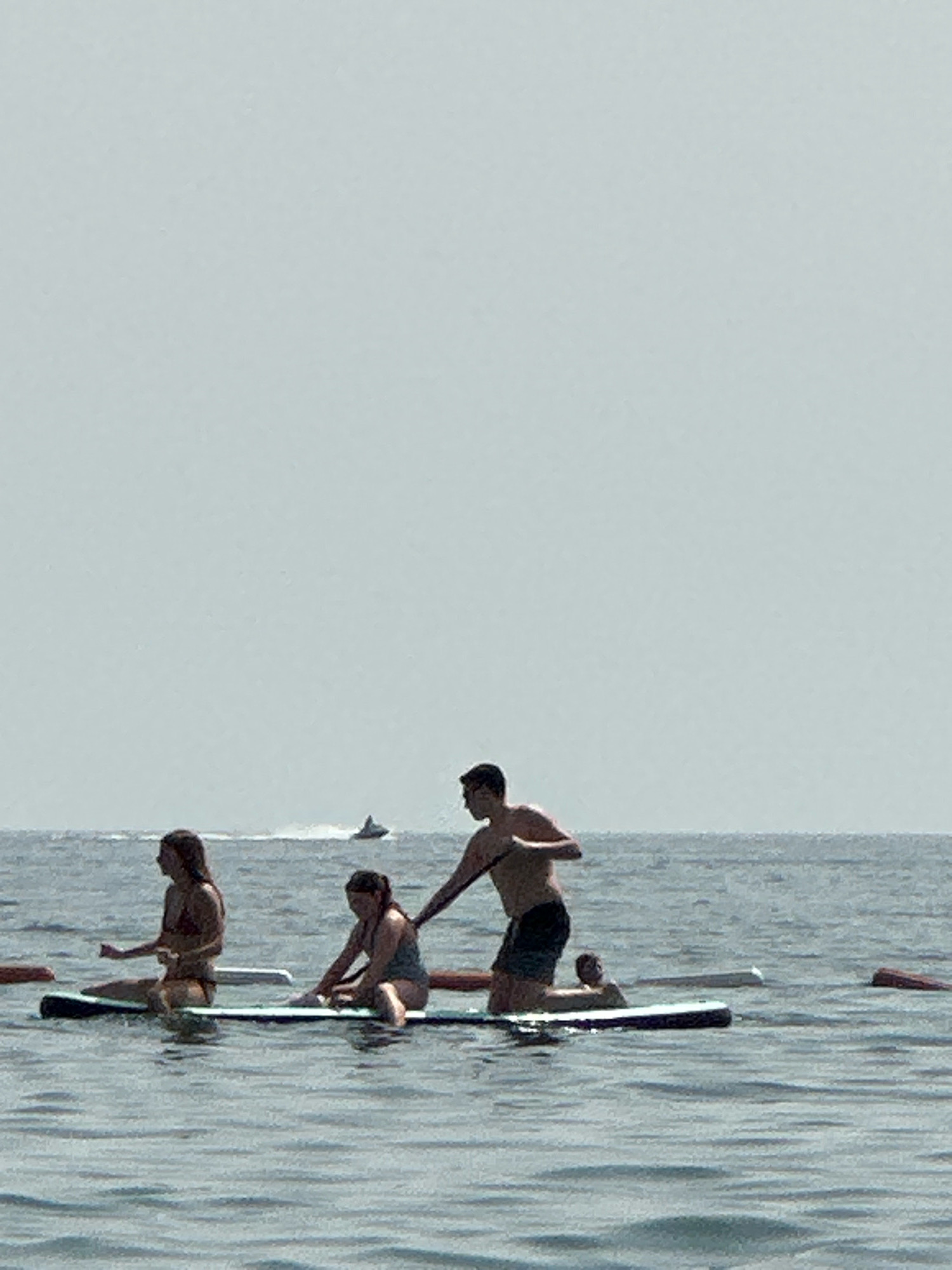 The image shows a group of people on paddleboards in the water. In the foreground, there are three individuals on separate paddleboards. A woman in a bikini is on the left paddleboard, sitting with her legs crossed. Next to her, on another paddleboard, a woman in a swimsuit is kneeling, and a man in swim trunks is kneeling behind her, reaching towards her. Further to the right, on a third paddleboard, another person is visible, partially submerged in the water. In the background, the sea extends to the horizon under a pale, overcast sky. A jet ski is visible in the distance. The water is calm with small ripples. The overall scene depicts recreational activity on the water, likely in a coastal or lake environment. There is no information in the image that specifically places the location in Boreti, Montenegro.