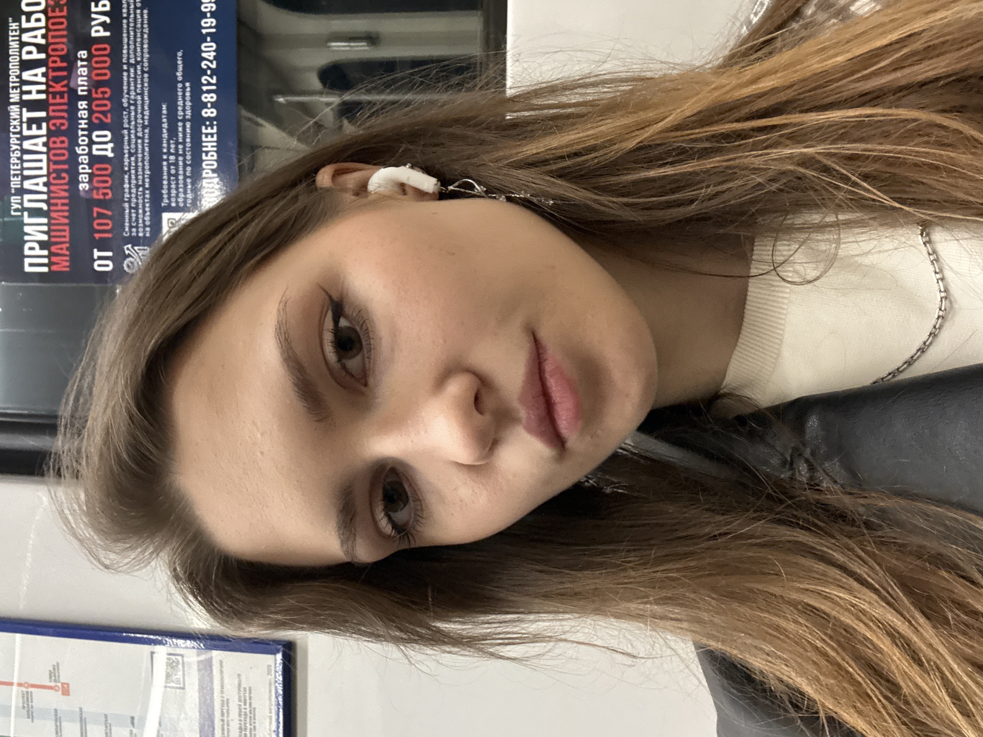 A close-up, horizontally oriented image features a young woman with long, light brown hair looking towards the camera. She has visible dark eyeliner around her eyes and slightly parted lips. A white earbud is seen in her right ear, and a silver chain necklace hangs around her neck. She appears to be wearing a white ribbed top and a black outer garment, possibly a jacket. Her expression is neutral.
The background primarily consists of a blue poster or sign with white and red text, and a white surface to the left. The sign is partially obscured by the woman's head and hair.
Visible text on the sign, written in Russian, indicates it is a job advertisement from "ГУП 'ПЕТЕРБУРГСКИЙ МЕТРОПОЛИТЕН'" (State Unitary Enterprise "Saint Petersburg Metro"). The ad invites "МАШИНИСТОВ ЭЛЕКТРОПОЕЗДОВ" (train operators) to work, listing a salary range of "ОТ 107 500 ДО 205 000 РУБ" (From 107,500 to 205,000 rubles). Benefits like a shift schedule, career growth, training, social guarantees, early retirement, and medical support are mentioned. Candidate requirements include being over 18 years old, having at least a secondary general education, and being in good health. A contact phone number, 8-812-240-19-95, is also provided. The setting strongly suggests the woman is in a Saint Petersburg Metro environment, likely inside a train car or station, with the poster serving as background information.