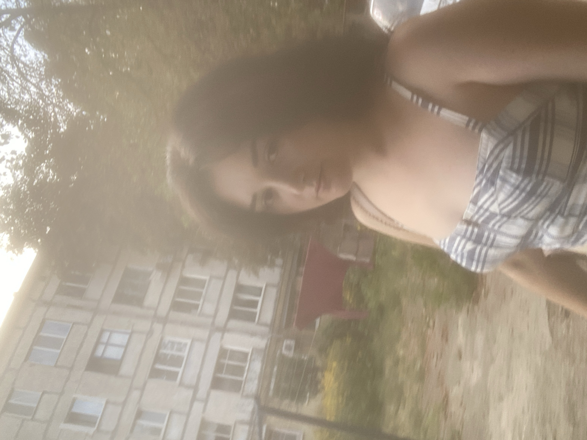 An out-of-focus, medium shot captures a young woman with dark, wavy hair and fair skin, wearing a plaid sundress, standing outdoors in front of a multi-story apartment building. The building, likely located in Poltava, Ukraine, has multiple windows and balconies. Greenery, including trees and bushes, surrounds the building and the woman. A red object, possibly a tarp or piece of fabric, is visible on the ground behind her. The lighting suggests it is daytime, with some sunlight filtering through the trees, creating a hazy effect. The woman's facial expression is neutral, and she is looking slightly to her right. Her left arm is down by her side, and her right arm is not fully visible. The overall scene depicts a casual, everyday moment in an urban environment.