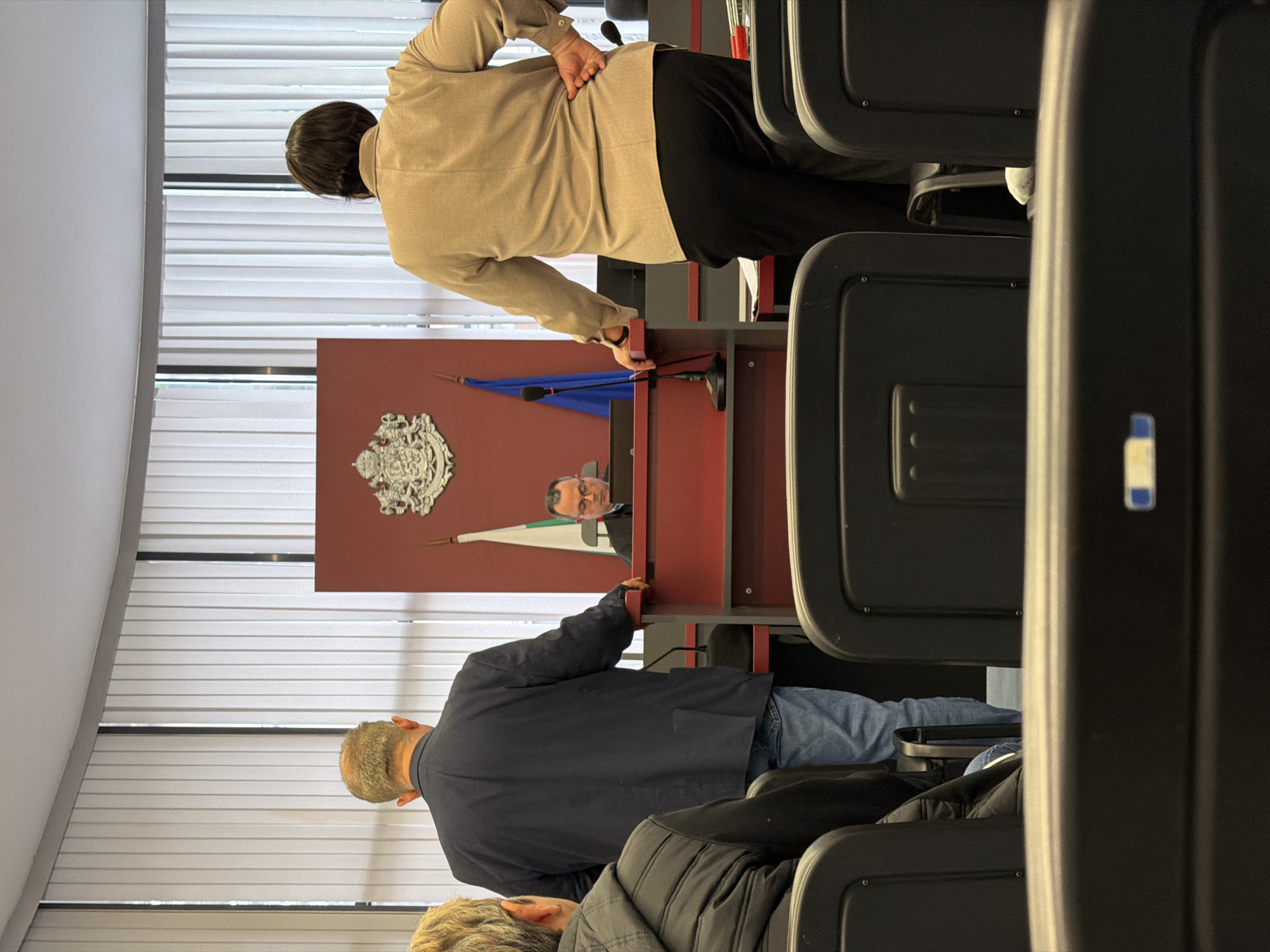 The image shows a meeting room in Plovdiv, Bulgaria. A man is sitting behind a desk with the Bulgarian coat of arms on the wall behind him, flanked by the Bulgarian and Plovdiv flags. Two people are standing at the desk, apparently listening to him. Other people are seated in rows of chairs in the foreground. The room has blinds on the windows. The environment seems to be a formal setting, likely a municipal or governmental office.