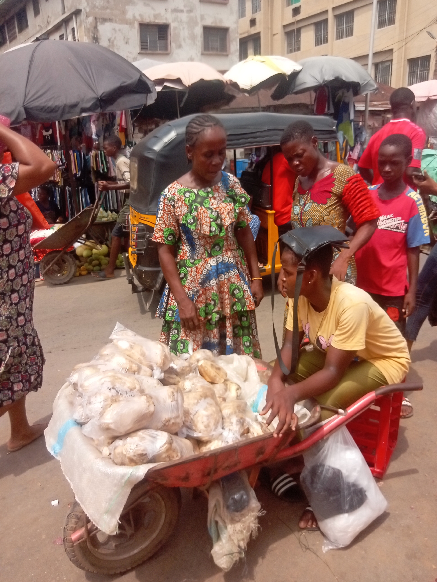 A bustling market scene in Onitsha, Nigeria, features a crowded street where a woman in a colorful patterned dress stands beside a red wheelbarrow filled with bags of produce, likely yams or cassava. A younger person wearing a yellow shirt and green pants sits on the wheelbarrow, possibly selling the produce. Another woman in a similar patterned dress and a child in a red t-shirt appear to be interacting with the vendor. The background shows market stalls with clothing for sale, covered by umbrellas, and an auto-rickshaw (keke) parked nearby, suggesting a commercial environment. Buildings are visible in the background. The scene conveys the typical energy and commerce of a Nigerian market.