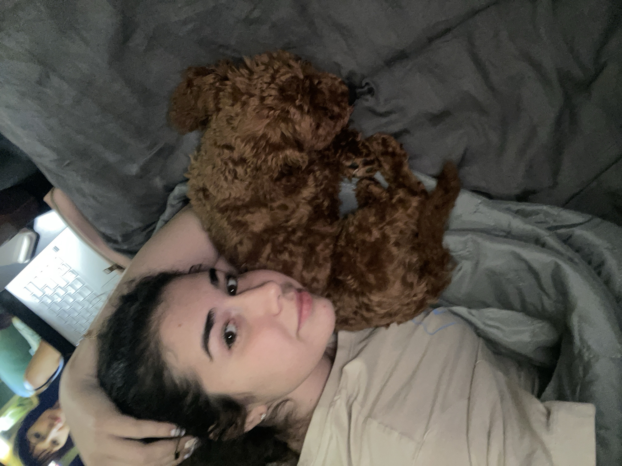 A young woman is lying on a bed with a brown poodle. The woman is fair-skinned with dark hair, and she is wearing a light-colored shirt. The poodle is curled up next to her. The bed has gray sheets and a gray comforter. A laptop is visible in the lower left corner of the image. The location is unknown, Sofia, Bulgaria is not evident.