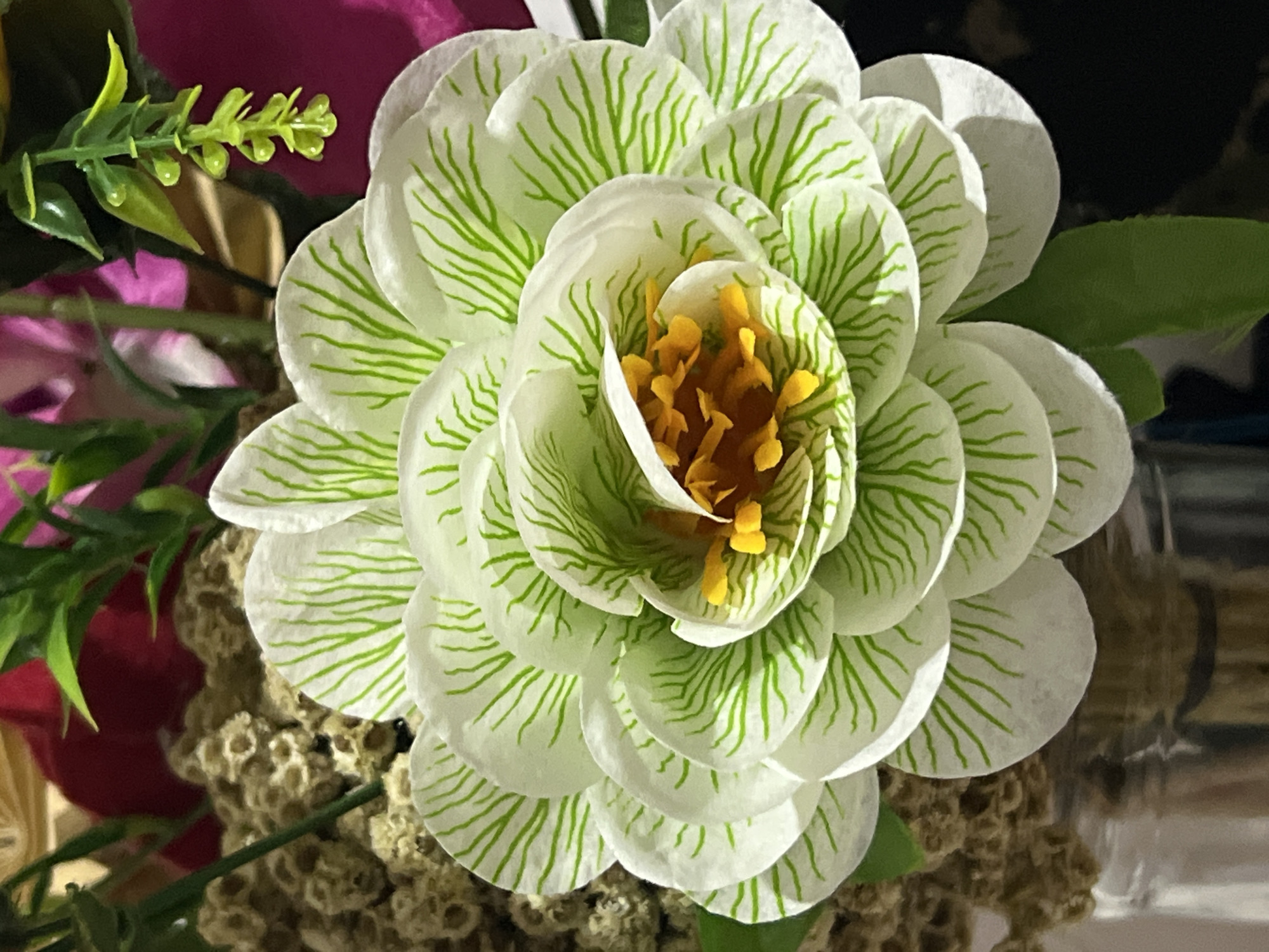 A close-up shot of a decorative artificial flower is presented. The flower has numerous white petals with thin green lines resembling veins. The center of the flower features yellow stamens. The flower is part of a bouquet that includes other floral elements, some appearing dried and textured, and green leaves. The bouquet is in a glass vase, the bottom of which is visible. The background is dark and out of focus. The scene is set in Denpasar, Indonesia.