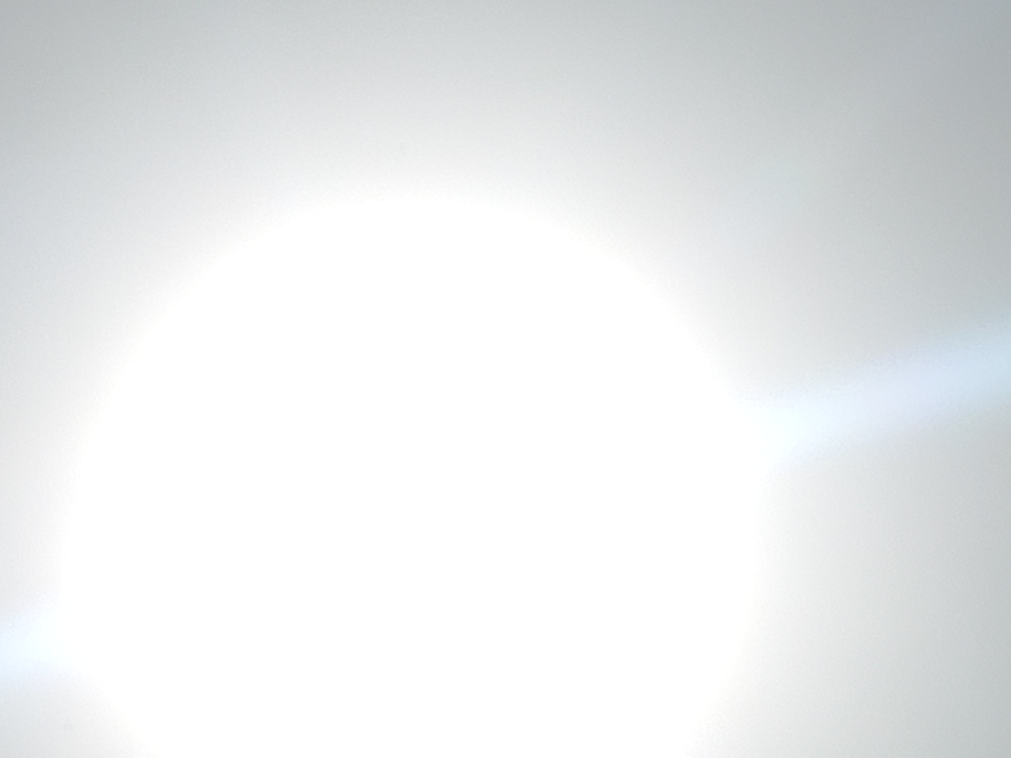 The image shows an out-of-focus, overexposed view, likely caused by direct sunlight or a strong light source aimed at the camera lens. The predominant color is white, with subtle gradations of gray suggesting a light-filled or reflective environment. No specific elements are discernible to provide location context related to Atani, Nigeria. No objects, actions, or people are visible.