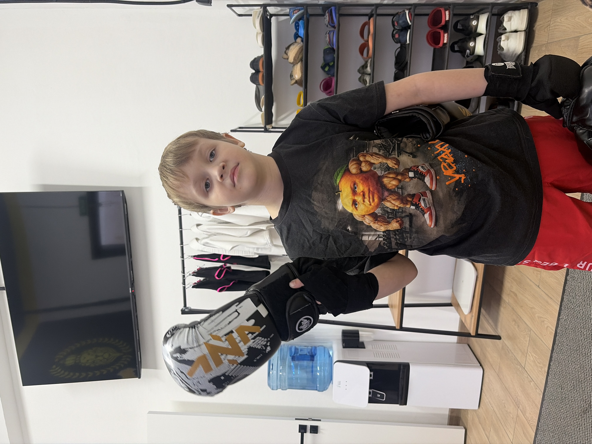 A young boy stands indoors, wearing boxing gloves and a black t-shirt with an image of a muscular orange with a human face. He's wearing red pants. Behind him is a shoe rack with multiple pairs of shoes, a clothing rack with clothes hanging on it, a water cooler with a blue water jug, and a TV mounted on the wall. The location is not discernible based on the provided image.