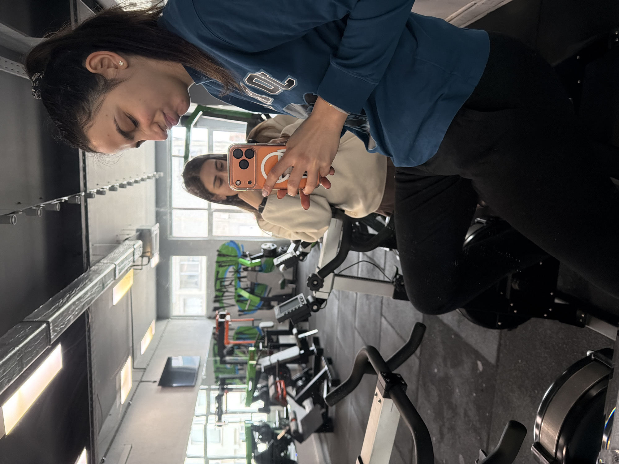 The image depicts two women in what appears to be a fitness center or gym. The woman in the foreground is wearing a blue sweatshirt and black pants, while the woman reflected in the mirror appears to be wearing a light-colored sweatshirt and black pants. The first woman is taking a selfie using an orange smartphone, capturing the reflection of the second woman in a mirror. The background includes exercise bikes and other gym equipment, suggesting the location is a fitness facility. The specific location in Sofia, Bulgaria, cannot be determined from the image alone, as there are no discernible landmarks or signs indicating its precise location.