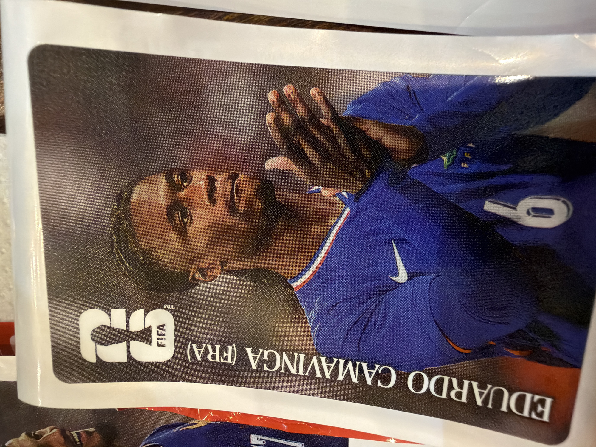 This is a close-up, eye-level, slightly angled photograph of a soccer trading card featuring Eduardo Camavinga. The card is positioned vertically and appears to be part of a collection, with white borders and a textured background. Camavinga is shown from the chest up, looking to his right with his hands clasped in front of him. He is wearing a blue soccer jersey with white trim around the neckline and a Nike swoosh logo on the right chest. The jersey also has three small green emblems with gold detailing on the left chest, and a large white number "6" on the lower left side of the image. A FIFA logo is visible in the upper left corner of the card. The text "EDUARDO CAMAVINGA (FRA)" is printed vertically along the left side of the card in white lettering. The background of the card is a dark, mottled texture, likely representing a stadium or a generic sports backdrop.