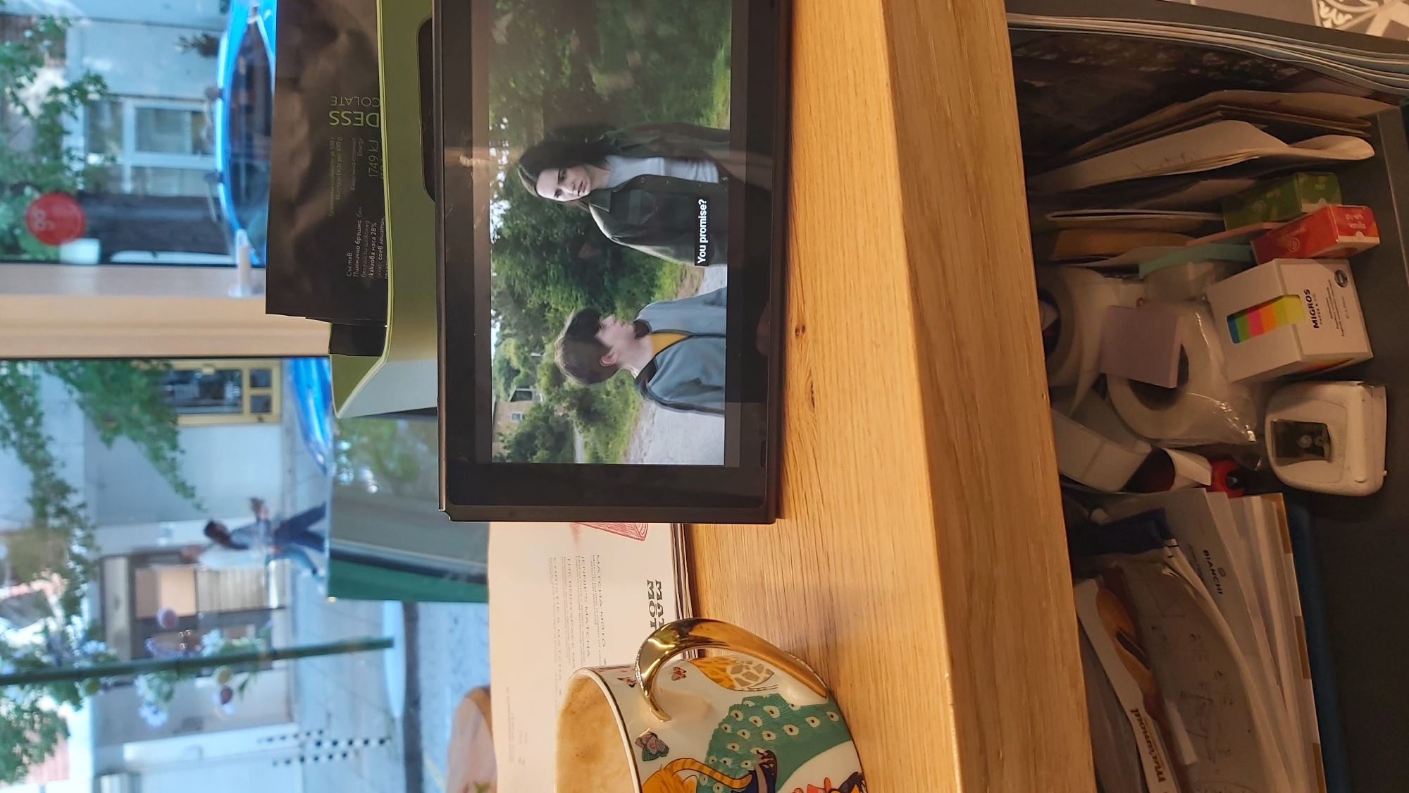 A high-angle indoor shot presents a scene cluttered with various objects, including a cup, a tablet displaying a movie, and a drawer overflowing with miscellaneous items.
The scene is likely set in a home or office environment. The foreground features a partly visible ornate cup, likely made of ceramic or porcelain, with a gold-colored handle. In the center is a tablet displaying a scene from a movie. On the screen, two people can be seen outdoors, one seemingly questioning the other. Text on the screen reads, "You promise?".
Behind the tablet is a partially opened wooden drawer filled with stationery items, tapes, a pen, and other office supplies. There are also some chocolate packets.
Through a window, a blurred outdoor scene shows buildings, trees, and people. This suggests the location could be in Sofia, Bulgaria.
