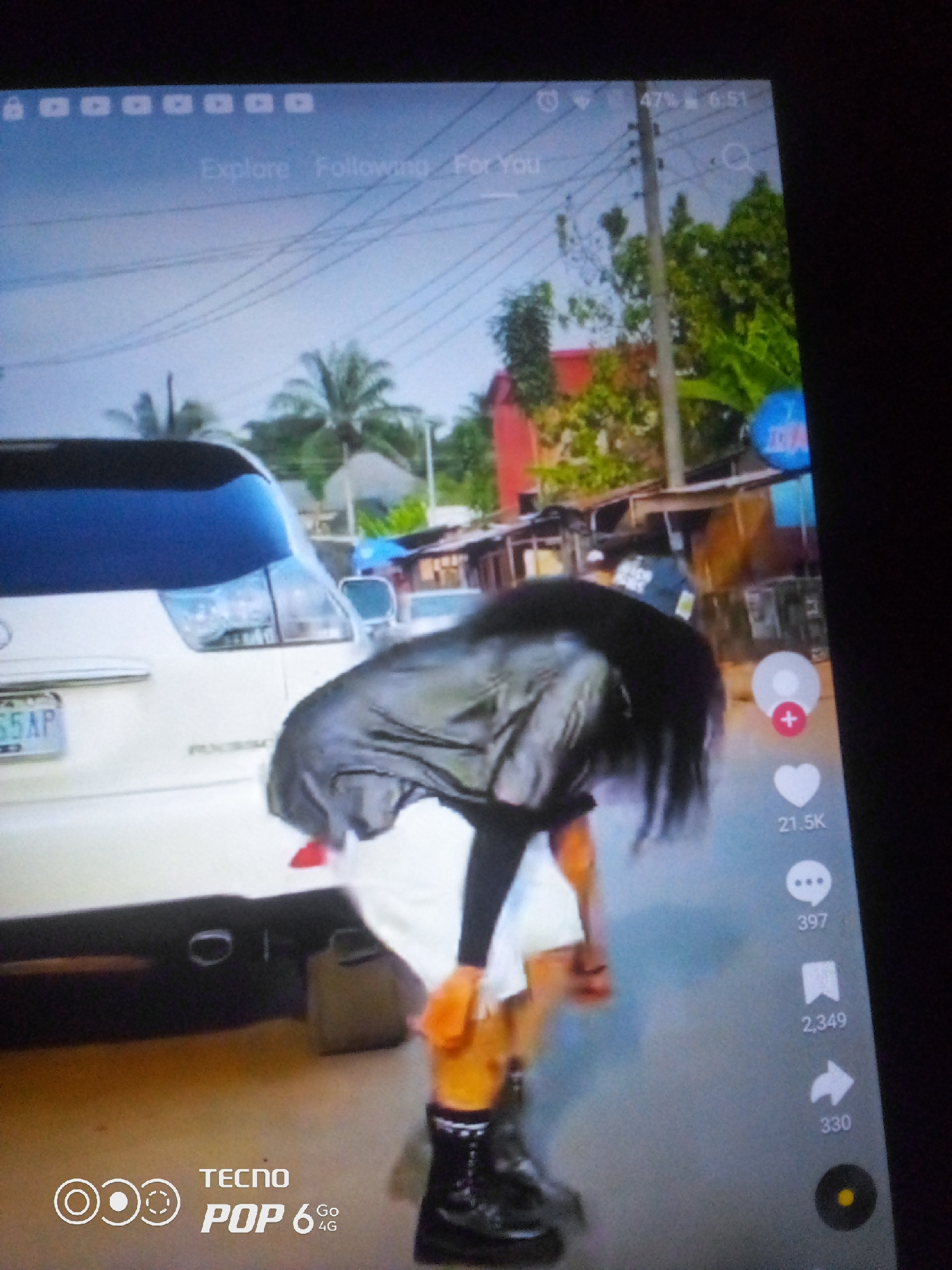 The image captures a low-angle shot of a person wearing black boots and a black jacket, with long dark hair obscuring their face. They appear to be in the process of bending over. In the background, a white SUV with a license plate reading "S5AP" is parked. The scene appears to be an outdoor environment, possibly a street, with trees and some buildings visible. The overall impression is that of a social media video being viewed on a device, as indicated by the on-screen icons and text such as "Explore," "Following," "For You," and engagement metrics like "21.5K" and "397." There is no visual information in the image to confirm the location in the city of Howayi, Nigeria.