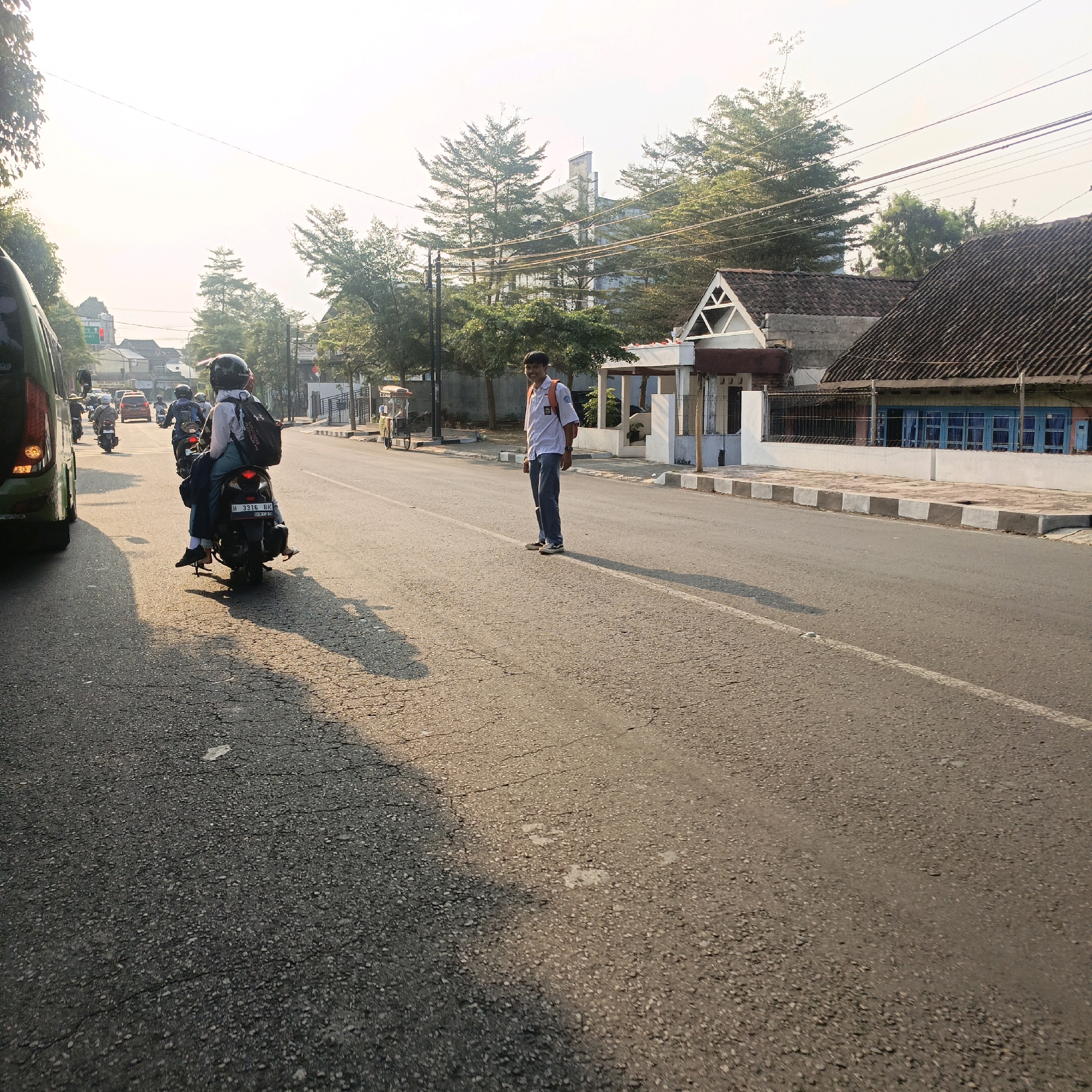 The image depicts a street scene in Salatiga, Indonesia, with a focus on road traffic and a young man standing by the roadside. In the foreground, a motorcycle with a rider wearing a black helmet and a backpack is moving away from the camera. The motorcycle has a license plate that reads "H 3316 BK". To the left, the rear of a green bus is visible, also in motion. Further down the road, several other motorcycles and a red car can be seen.
A young man, dressed in a white school uniform shirt with an orange backpack, stands facing the road, looking towards the camera with a smile. He is wearing blue jeans and dark shoes. Behind him, trees line the street, and buildings with tiled roofs are visible, including one with a triangular architectural feature. The sky is bright and hazy, suggesting it might be morning or late afternoon. The road surface is asphalt, showing signs of wear and tear with cracks and patches. The scene appears to be an urban or semi-urban environment, characteristic of Indonesian cities.