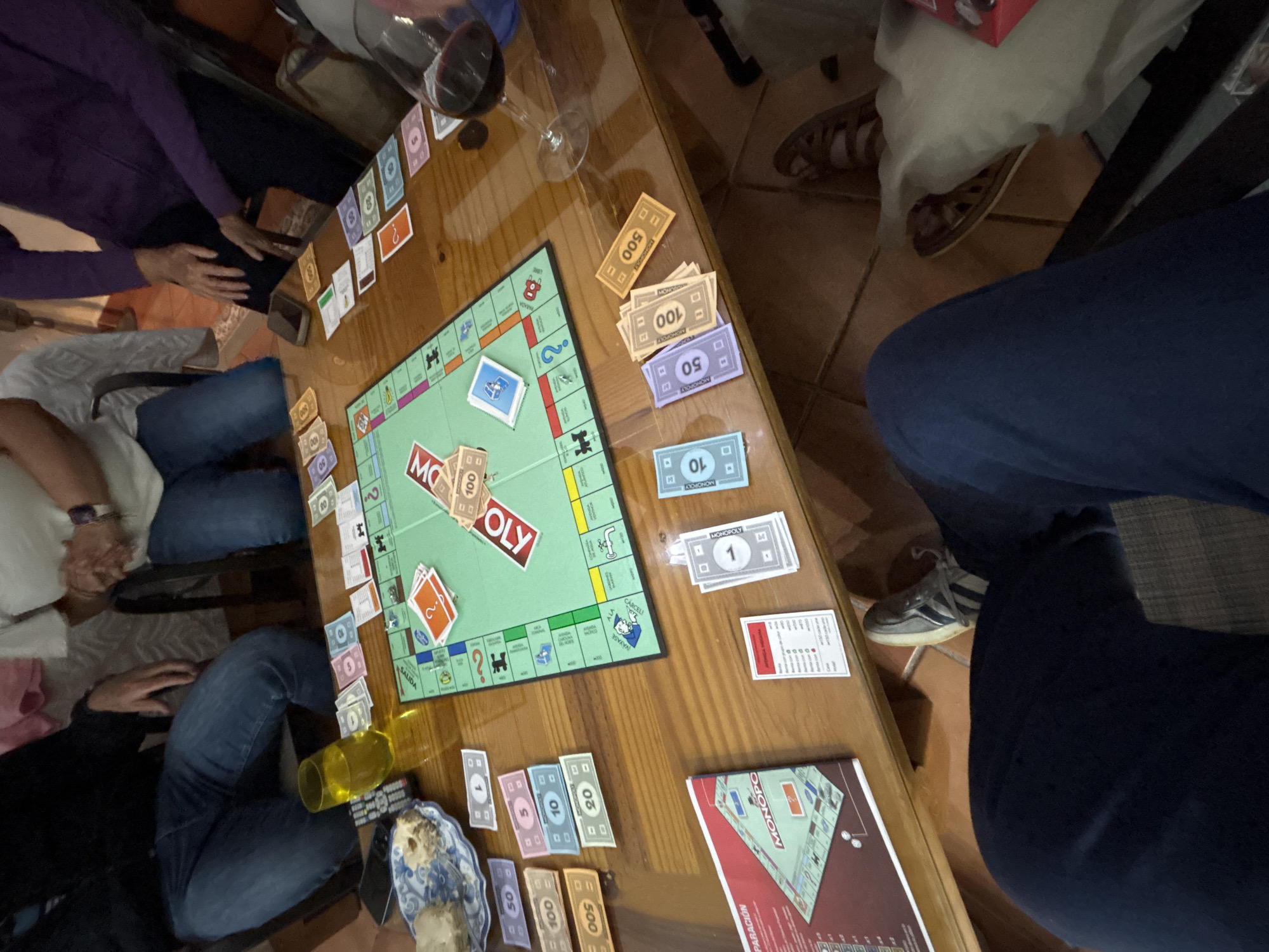 The image shows a group of people playing Monopoly around a wooden table. Monopoly game elements, including the board, money, property cards, and rulebook, are visible on the table. There are people seated around the table, engaged in the game. A glass of red wine is also present. The location is not discernible from the image.