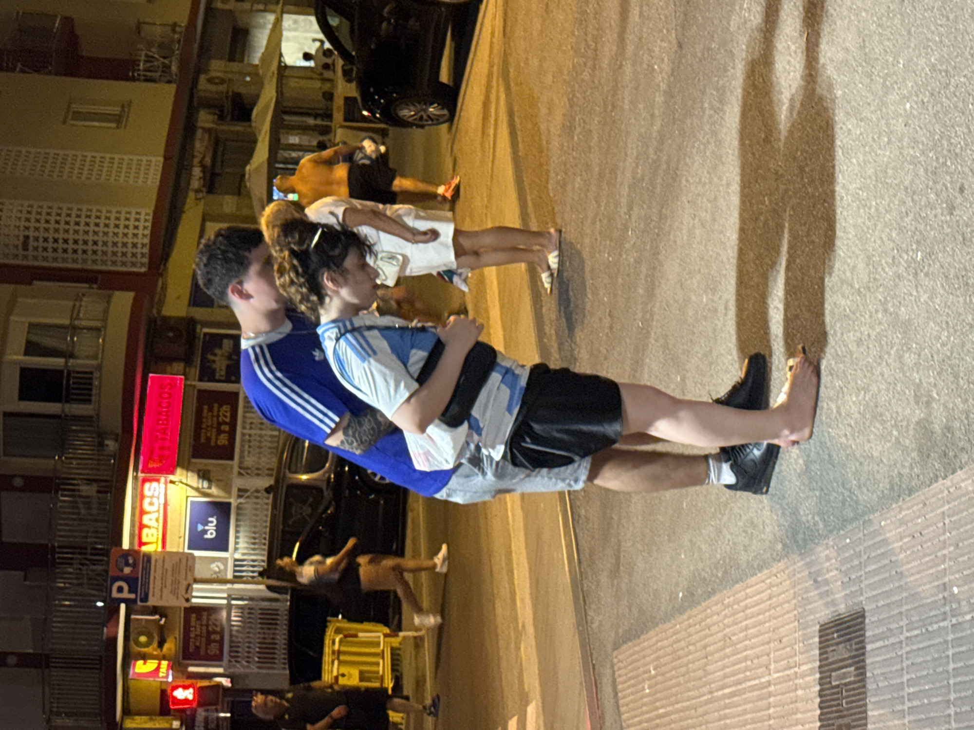 The image depicts a street scene in Blanes, Spain, at night. In the foreground, a man and a woman are embracing. The man, wearing a blue and white striped shirt and grey shorts, has his arm around the woman. The woman, also in a blue and white striped top and black shorts, is looking down. Both are barefoot. Behind them, other people are visible, including a man in a dark shirt on the left and a man with his back to the camera on the right. The background shows buildings with illuminated signs, including "ABACS" and "TABACOS", as well as a "blu." logo. A black car is parked on the right side of the frame. The ground appears to be asphalt, with a crosswalk marked in the center of the image. The lighting suggests it is evening or night.
