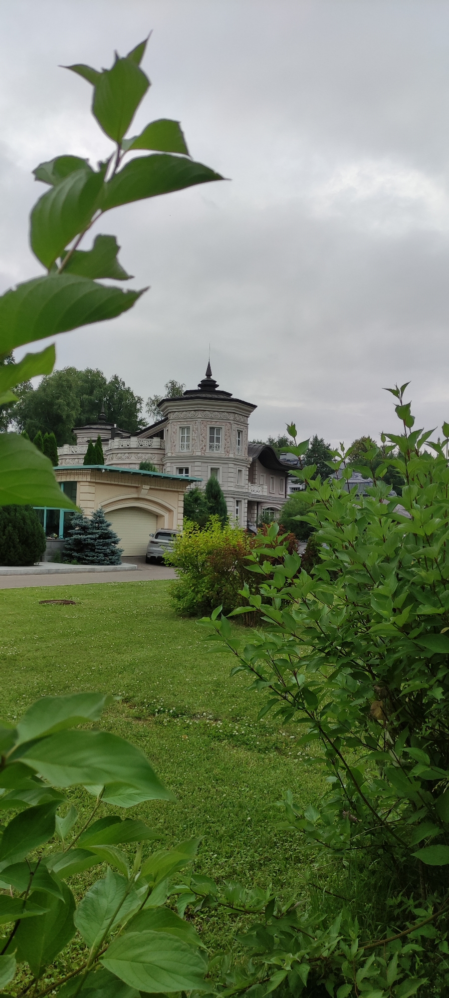 A grey, overcast sky looms above a large, ornate mansion with a circular tower in the city of Ангелово Клубная Резиденция, Russia. The mansion, painted in a light, neutral color, features decorative stucco work, multiple windows, and dark, conical spires atop its towers. In front of the mansion is a well-maintained green lawn dotted with small white flowers. To the left of the lawn, a large garage with a beige, arched door is visible, with a silver car parked outside it. Several evergreen trees, including a conical blue spruce, are planted in the vicinity of the garage and the mansion. Lush green foliage from nearby trees and bushes partially obscures the view of the mansion, framing the scene from the foreground. The overall impression is one of a grand, somewhat secluded residence on a cloudy day.