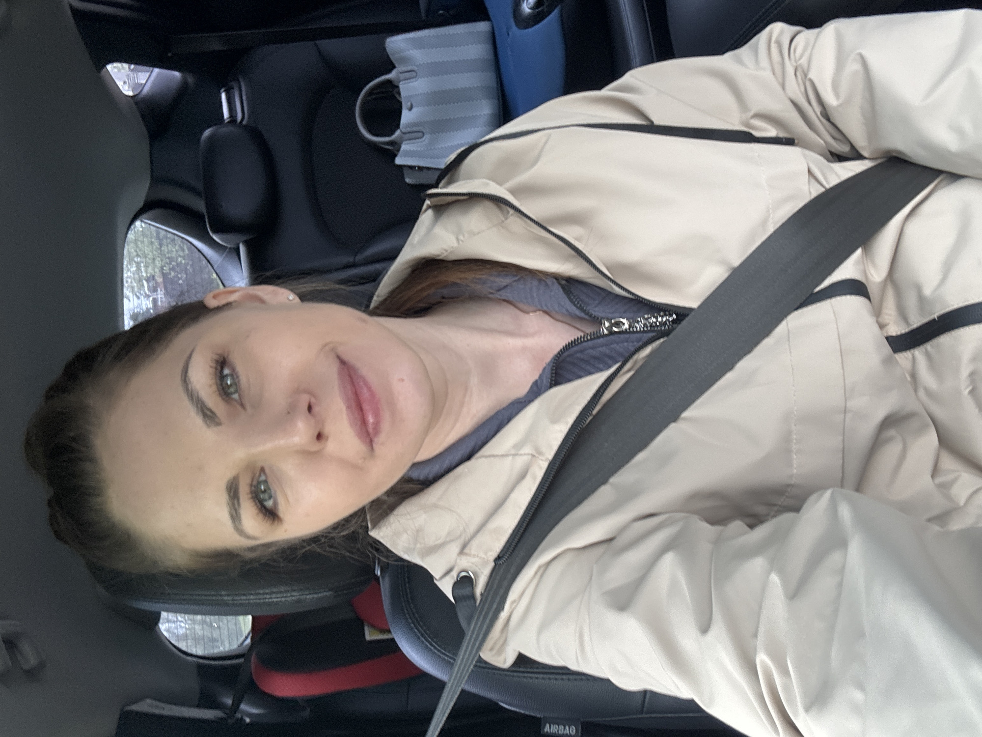 A woman is seated in the driver's seat of a car, wearing a seatbelt and a light tan jacket. The car is likely located in Раздолье, Russia, as that location was provided with the request. The car's interior is visible, including the steering wheel, dashboard, and a child's car seat in the back. The woman is looking at the camera.
