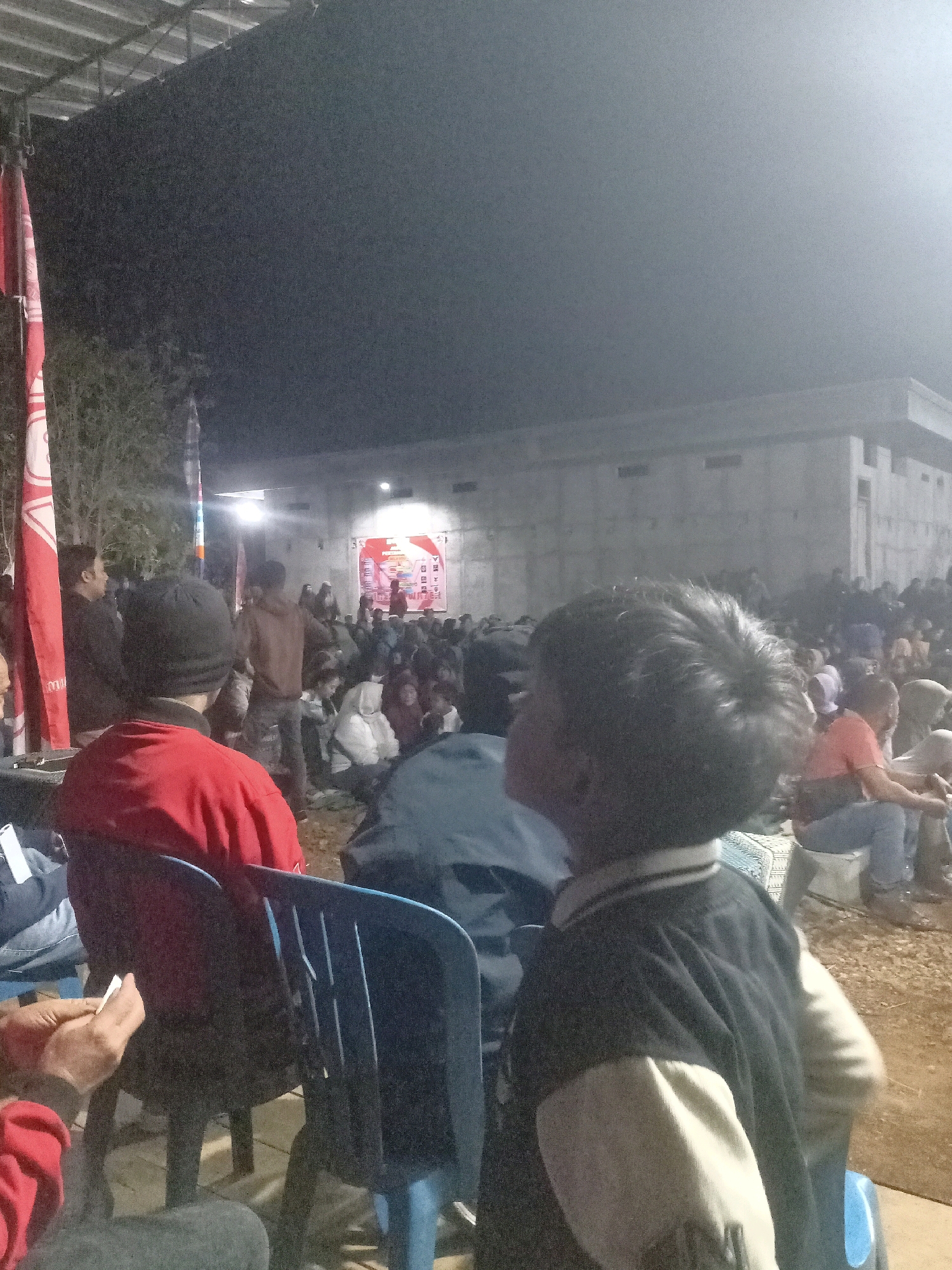 In Tasikhargo, Indonesia, an outdoor evening gathering is depicted. The foreground is dominated by a young person in a black and tan jacket, facing away from the camera and towards a crowd. In the middle ground, an individual in a red jacket is seated in a blue chair, also facing the gathering. To the left, two red flags with white markings are visible, and a white banner is draped. The background features a large crowd of people seated on the ground, some under a canopy, with a building structure behind them. The scene is illuminated by artificial lights, suggesting an event or performance is taking place. The overall atmosphere is communal, with people gathered to watch something.