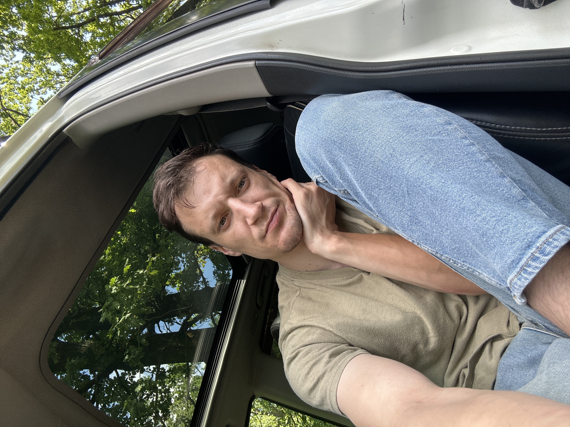 A medium shot captures a man sitting in the driver's seat of a white vehicle, with his left arm resting on his bent knee and his right hand supporting his chin. He is wearing a tan t-shirt and light blue jeans. The car's sunroof is open, revealing a canopy of green trees and a clear blue sky above. The vehicle appears to be parked on a dirt road or in a natural setting, possibly in the village of Makiv, Ukraine, given the surrounding foliage and rustic environment. The man is looking directly at the camera with a neutral expression.