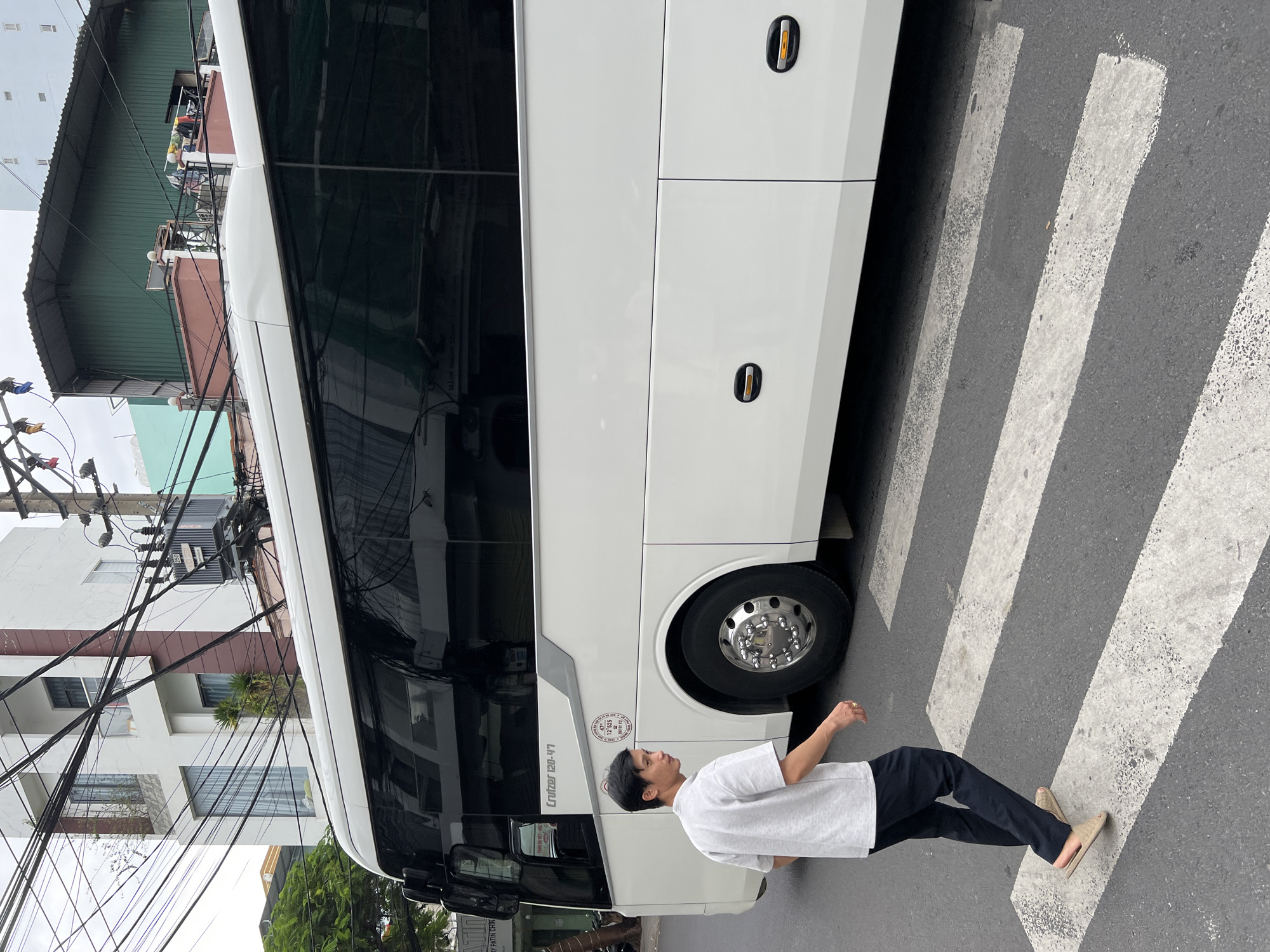 A medium shot captures a man crossing a pedestrian walk in Nha Trang, Vietnam, with a white bus to his right. The man, wearing a light grey t-shirt and dark trousers, walks from left to right, his gaze directed away from the camera. He is wearing beige slides. The bus, a prominent white vehicle with tinted windows, occupies the right half of the frame, its front portion obscured. The pedestrian crossing is clearly marked with white stripes on asphalt. In the background, urban elements such as buildings with balconies and windows, overhead electrical wires, and a utility pole are visible. The sky is overcast. The scene appears to be a street in a city with typical urban infrastructure.