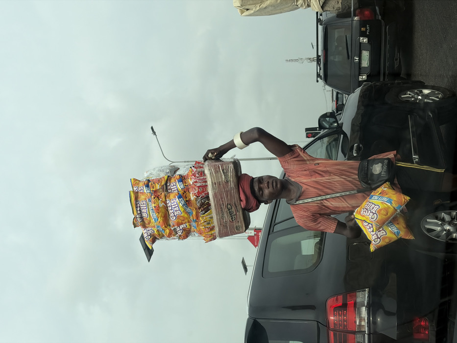 A male street vendor stands amidst stationary traffic on a road in Ikate, Nigeria, under an overcast sky. He is dark-complexioned, wearing a red and black striped shirt, black shorts, and a red head wrap. A white bangle adorns his left wrist, and a black "iPhone" branded crossbody bag hangs over his right shoulder. He carries a tall, rectangular stack of merchandise on his head, primarily consisting of large yellow bags labeled "Cheese Balls PARTY PACK," with smaller snack bags beneath, all secured by clear plastic. In his left hand, he holds two additional "Cheese Balls PARTY PACK" bags. His right arm is raised, with his hand near the top of the merchandise stack. To his left is a dark-colored SUV, likely a Toyota Land Cruiser, displaying its rear lights. Behind it and to the right, a black Honda Pilot is visible. A street light pole and a red traffic signal are present in the background.