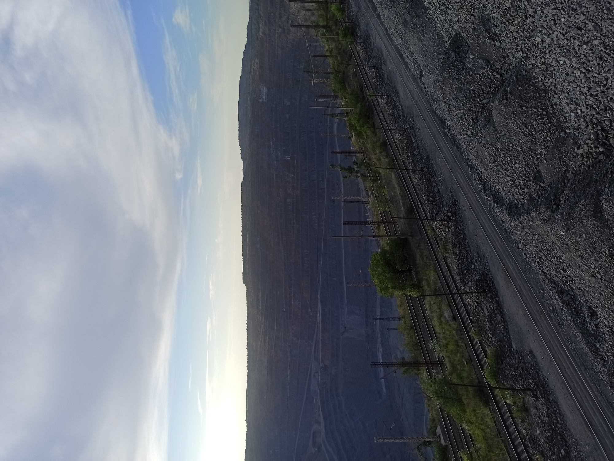 The image depicts a massive open-pit mine, likely a quarry, with extensive layered excavations visible on a steep mountainside. The scale of the operation is evident in the sheer depth and width of the cut. Multiple railway tracks run alongside the mine, suggesting the transportation of extracted materials. These tracks are bordered by areas of loose rock and sparse vegetation, including some trees. The sky above is partly cloudy, with wispy white clouds against a bright blue backdrop, indicating fair weather. The lighting suggests it is either early morning or late afternoon, with a gentle, diffused sunlight. There is no visible text in the image. The overall scene conveys a sense of industrial activity and the raw, powerful force of nature being reshaped by human endeavor. The location context of Kryvyi Rih, Ukraine, known for its iron ore deposits, further reinforces the interpretation of this as a mining operation.