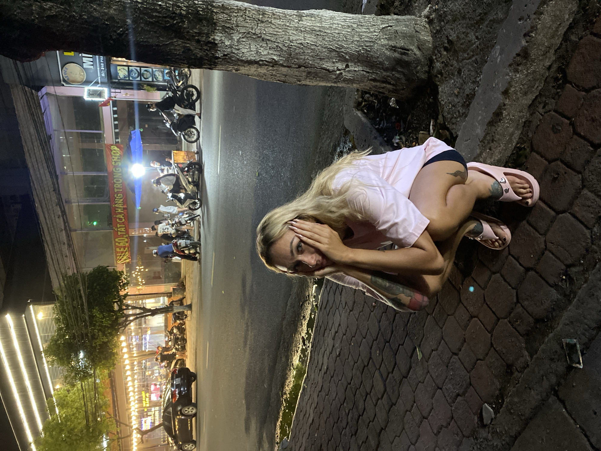 A woman with blonde hair is crouching near a tree trunk at night. She is wearing a light pink t-shirt, dark shorts, and pink sandals. Her hands are placed on her face. The background includes a street with parked motorbikes and a building with lights and signs in Vietnamese. The sign reads "LE 60. TAT CA HANG TRONG SHOP." The scene appears to be in Đồng Nai, Vietnam.