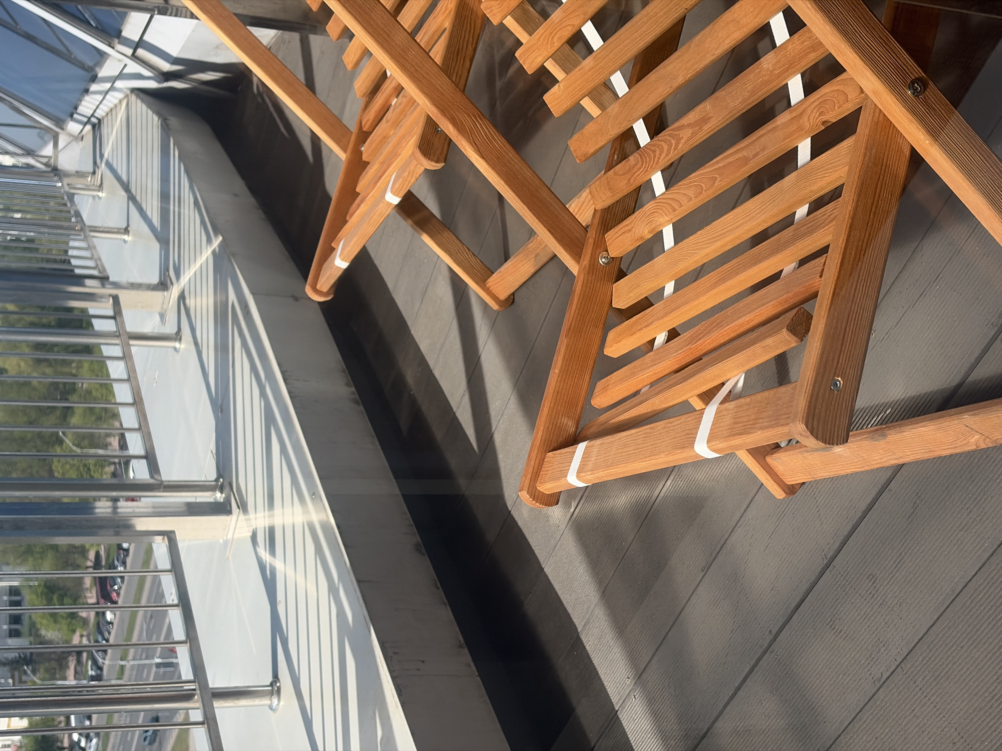 On a balcony in Minsk, Belarus, two foldable wooden chairs with white webbing are angled towards each other on a gray wooden-planked surface. The balcony has a metal railing. Reflected in the glass panel is a view of trees, cars, and city streets. The light source is illuminating the scene from the right.