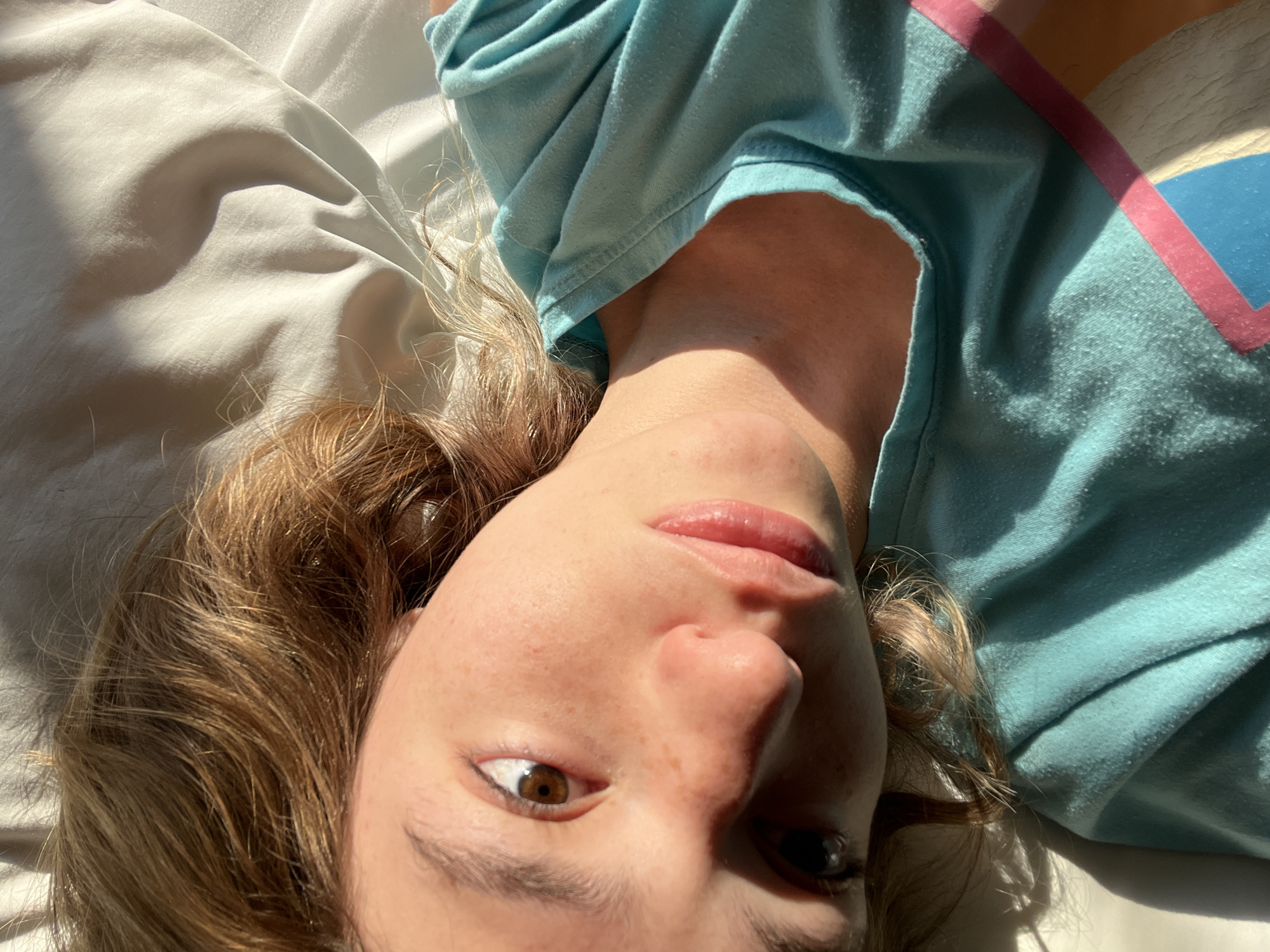 A person is lying down on a white surface, possibly a bed. The individual is wearing a light blue shirt with a pink stripe along the collar. Their hair is light brown. They have fair skin and brown eyes. The sunlight is casting strong shadows across their face and shirt. There is no information to identify any location in San Bernardino Tlaxcalancingo, Mexico.
