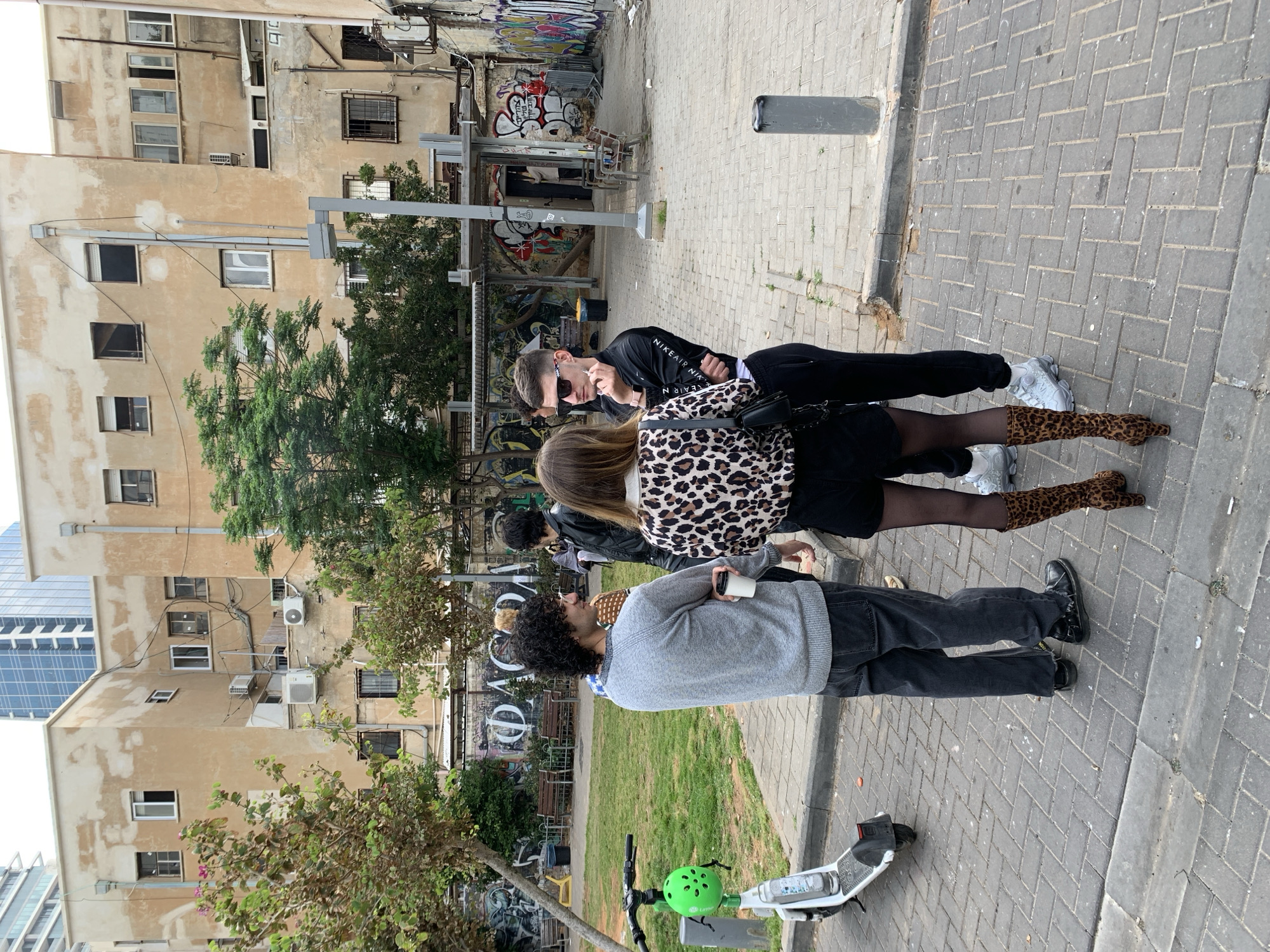 The image captures a scene on a paved sidewalk in Tel-Aviv, Israel. Three people are visible: a male wearing sunglasses and a black Nike Air jacket, a female with a leopard print jacket, and another individual with curly hair wearing a gray sweater. The first two people are standing on the sidewalk in front of a bus station. The third person is holding a disposable cup and standing on the sidewalk next to a small patch of grass and a shared electric scooter with a green helmet attached to its handlebars. Behind the group, buildings with graffiti-covered walls and aged facades are visible. A modern skyscraper is partially visible in the distance.