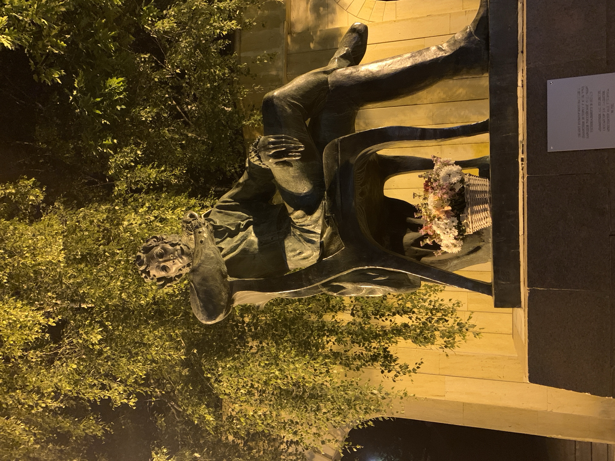 The image depicts a bronze statue of a man seated on a chair, located outdoors in the city of Unknown, Unknown, at night. The statue portrays a man with curly hair, dressed in a jacket, trousers, and boots, seated with his legs crossed at the ankles. His right arm rests on the armrest of the chair, and his left hand is raised towards the chest area. The statue is mounted on a dark, multi-tiered stone pedestal. A wicker basket containing various flowers, primarily white, pink, and red, is placed on the upper tier of the pedestal, to the right of the statue's legs. A rectangular silver plaque with text is affixed to the front of the lower pedestal tier. Behind and above the statue, dense green tree foliage is brightly illuminated by artificial light, giving the leaves a yellowish-green appearance against a dark background. A light-colored stone or concrete wall, featuring an archway on the left side, is partially visible behind the statue. The scene is lit by warm, artificial light, casting shadows and illuminating the statue and immediate surroundings while the distant background remains dark.