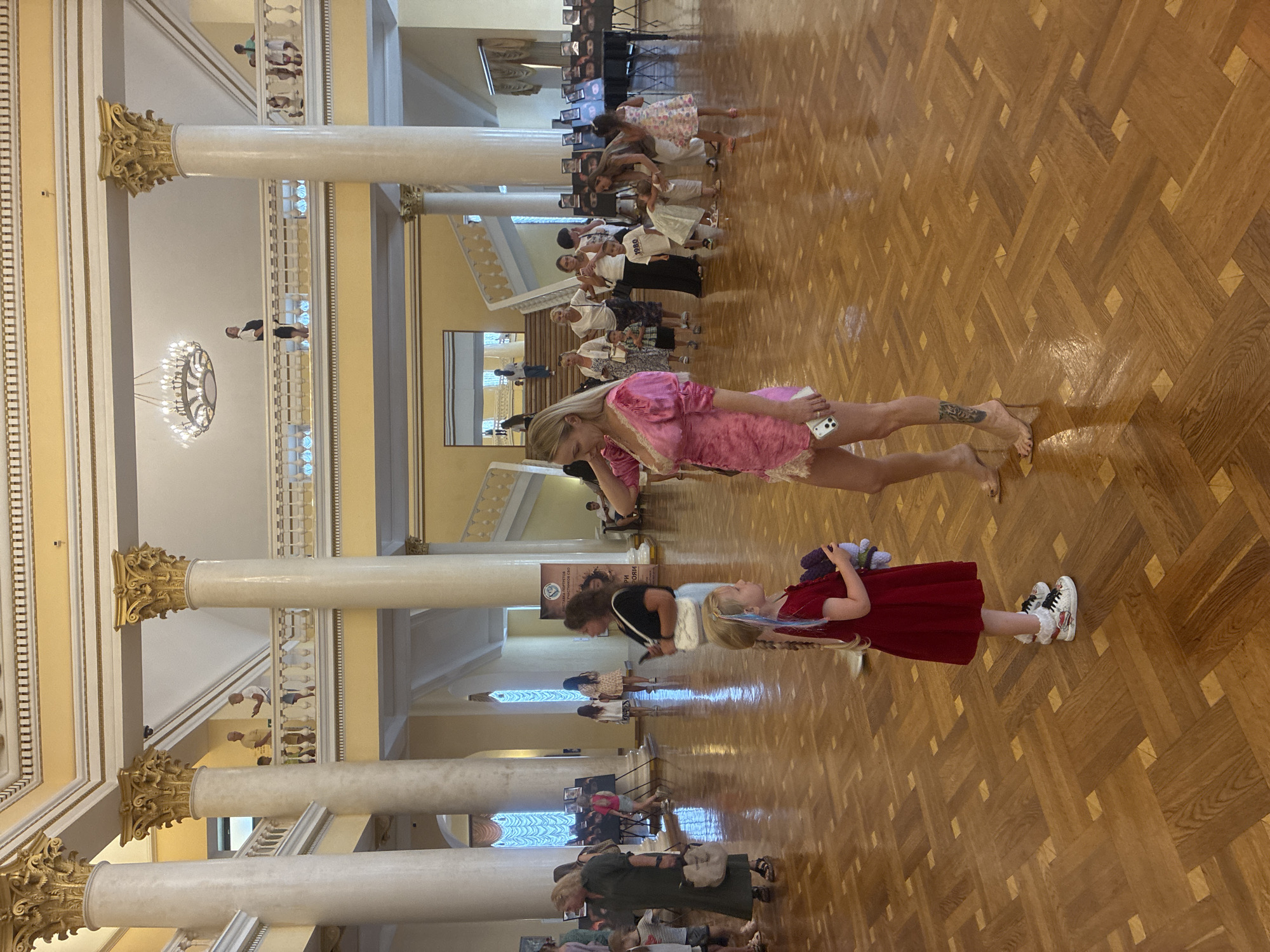 In a grand hall with polished wooden floors and large columns, two women and a young girl are the central focus. One woman, wearing a pink dress and high heels, is holding a phone. She is interacting with a young girl in a red dress, who is holding a stuffed toy. Other people are visible in the background, some walking and others observing an exhibition of photographs on tables. The architecture suggests a public building, potentially a museum or cultural center, given the exhibition. The image captures a moment of interaction amidst a gathering. The location is a hall in the city of Sochi, Russia.