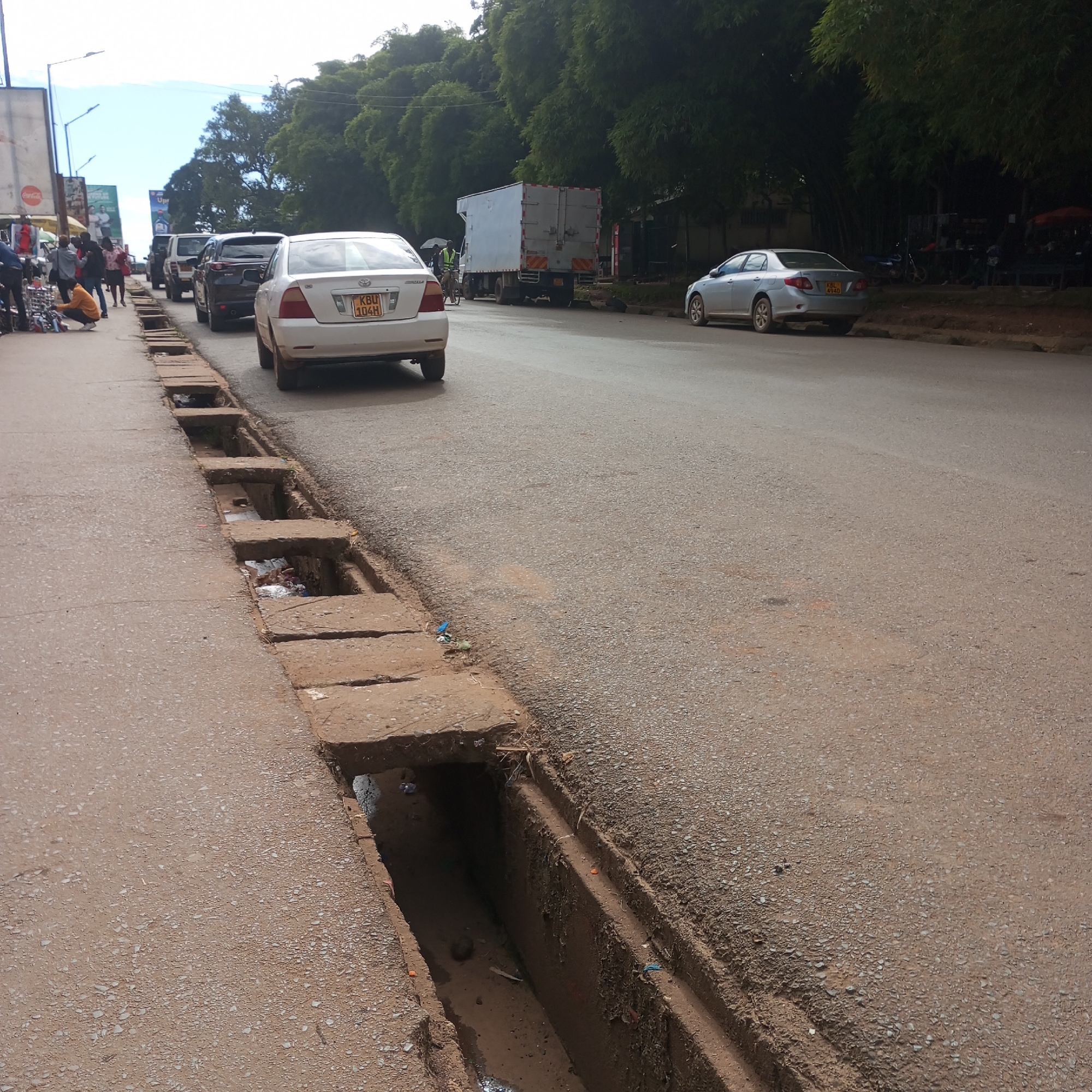 The image shows a street scene in Sheywe ward, Kenya. A drainage ditch runs along the left side of the asphalt road, covered intermittently with concrete slabs. The ditch contains debris and stagnant water. Several cars are parked or moving along the road, including a white sedan with license plate KBU 104H. A truck is also visible further down the street. People are present on the sidewalk on the left side of the road, near a Coca-Cola sign. Trees line the background.