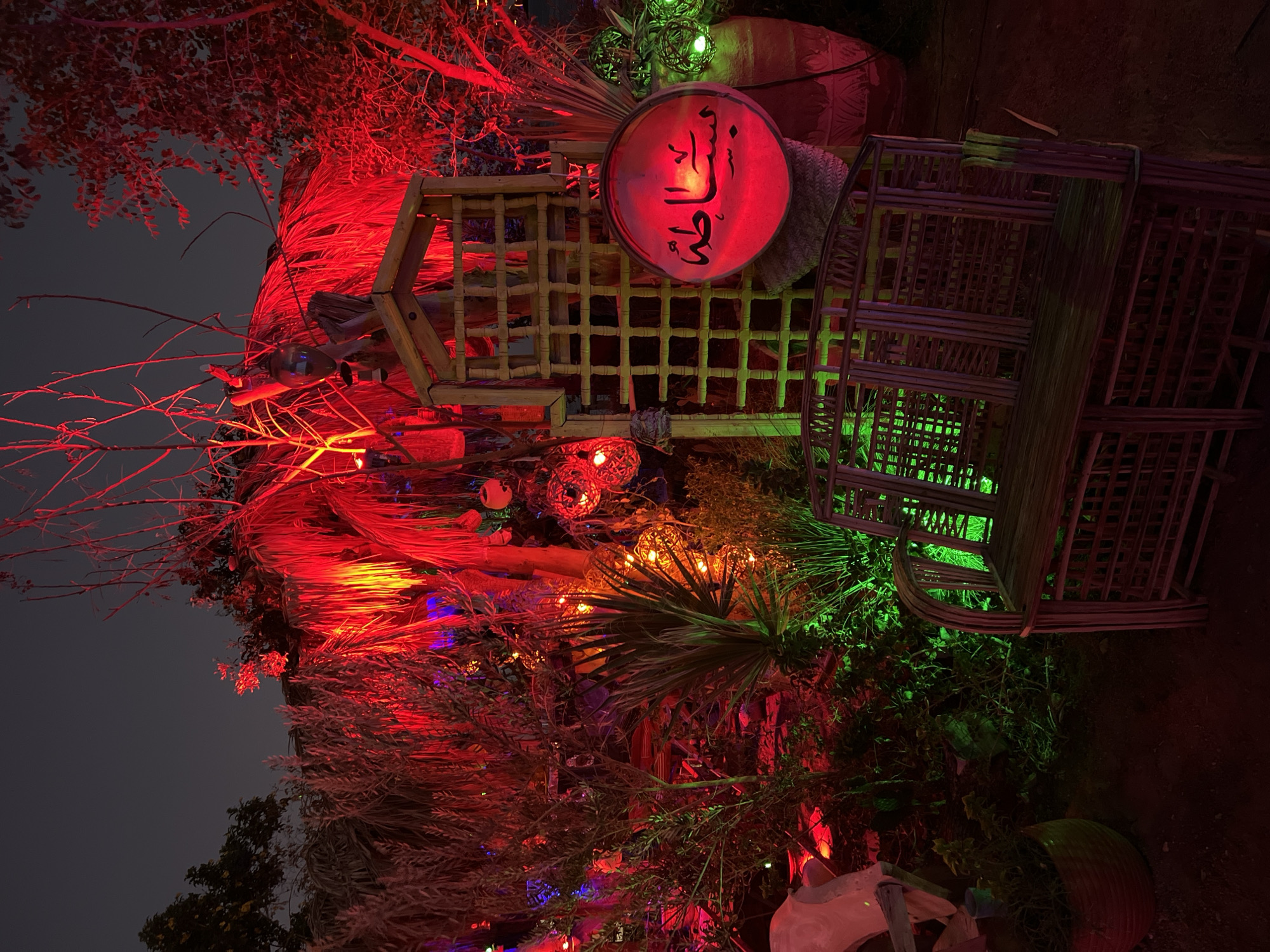 This image depicts an outdoor garden scene in Sharm El Sheikh, Egypt, illuminated by vibrant red and green lights. The setting appears to be a decorative display, possibly for an event or celebration. In the foreground, a rustic wooden bench made of woven reeds sits on dark soil. Behind the bench, a wooden lattice structure is visible, with a circular red object displaying Arabic script hanging to its right. The surrounding vegetation is heavily lit, with palm fronds and other plants appearing in shades of red and green. Several decorative spherical lanterns, some also lit with red and green lights, are scattered throughout the scene. In the background, more trees and foliage are visible, their colors intensified by the lighting. The overall impression is one of an atmospherically lit, festive outdoor space.