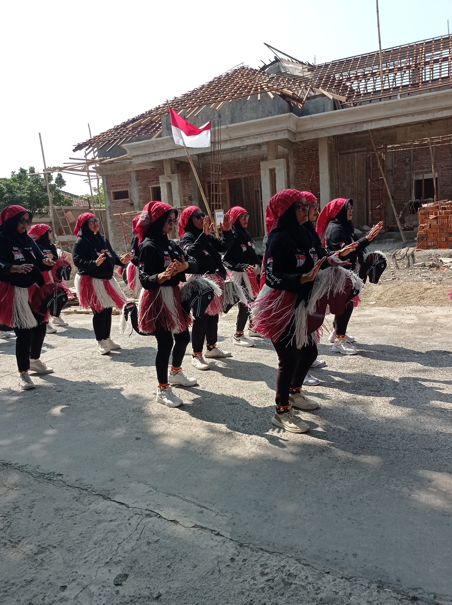 A group of women in traditional Indonesian attire, featuring red headscarves and white and red fringe skirts, are participating in a parade or performance. They are wearing black tops with white lettering. Some of the women are holding what appear to be miniature horse props made of dark material with white manes. The backdrop is a construction site, with exposed brick walls, scaffolding, and roofing in progress, suggesting a location in Maribaya, Indonesia. A red and white Indonesian flag is visible flying from a pole near the construction. The ground is paved and cracked, with shadows cast from the subjects and nearby trees. The women are in various stages of movement, with their hands raised and bodies positioned as if dancing or performing.
