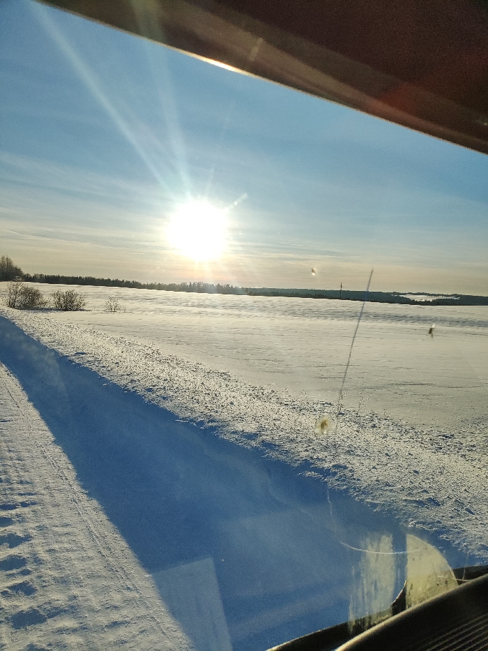 The image depicts a snow-covered winter landscape, viewed from inside a vehicle in the city of Малая Уса, Russia. The perspective is through a windshield or side window, with a dark brown vehicle frame visible at the top and a dashboard reflection in the lower right. A bright, unclouded sun is prominent in the upper left, casting strong rays across a clear blue sky with faint white clouds. The foreground and midground consist of expansive, flat snow-covered fields. A deep blue shadow extends diagonally from the lower left towards the center. Subtle tire tracks or disturbed snow are visible in the immediate left foreground. Sparse, leafless bushes dot the snow on the left side. A distant, dark treeline of what appears to be evergreen forest spans the horizon. Beyond this treeline on the far right, another snow-covered expanse is visible, possibly a frozen body of water or further fields, with a thin vertical dark object, possibly a utility pole, rising above the distant horizon. The vehicle's window exhibits multiple specks of dirt, smudges, and a thin vertical piece of debris in the mid-right portion, along with a prominent yellowish circular smudge in the lower right. No individuals or active subjects are present. The scene is brightly illuminated by direct sunlight.