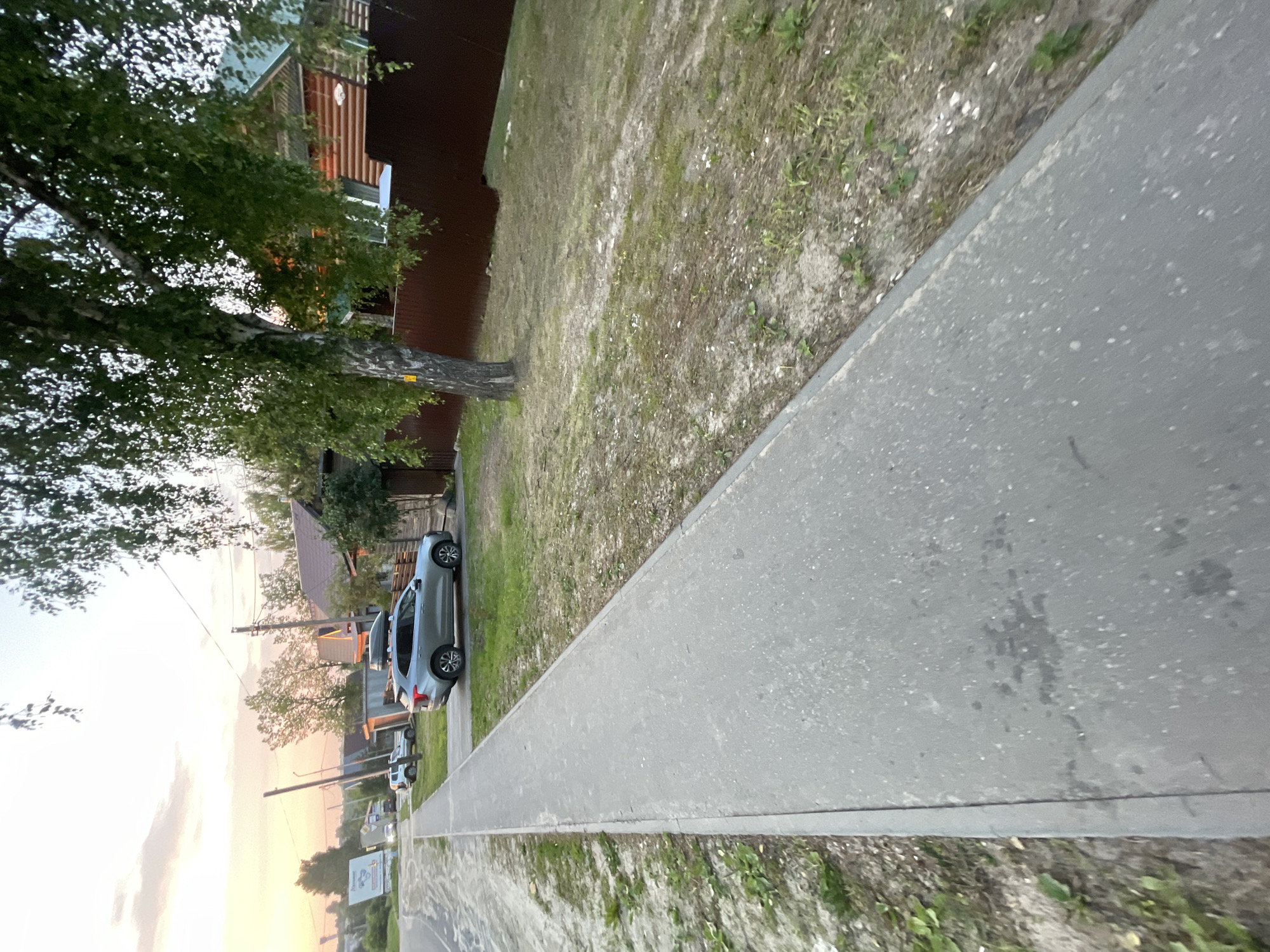 The image depicts a street scene in Krasnye Baki, Russia. In the foreground, a grey asphalt sidewalk runs diagonally from the bottom left towards the middle right of the frame. Adjacent to the sidewalk on the right is a patch of dry grass and dirt with scattered small green plants. Further to the right, a wooden house with a green roof and brown horizontal siding is visible, partially obscured by a tall, leafy tree with a grey-brown trunk. A dark brown fence runs along the property line. Parked on the grass area near the house is a silver SUV with a roof rack. In the background, a paved road continues, with other houses and trees lining the street. A large billboard with a white and green sign is visible on the left side of the road. The sky above is a soft pastel mix of orange, pink, and white, suggesting it is either sunrise or sunset. There are no people or animals visible in the scene.
