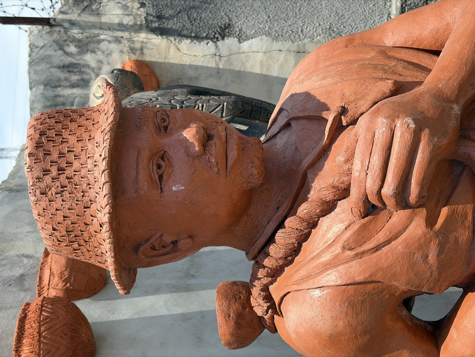 This image features a terracotta-colored statue of a male figure, positioned outdoors in Igboefon, Nigeria. The statue is depicted from the chest up, facing slightly right with its gaze directed downwards. The figure wears a woven-textured hat and has facial hair, including a mustache and short beard. A white smudge is present on the left side of its face, below the eye. A braided, thick strap extends over its right shoulder, securing a small, terracotta pot. The figure's hands are clasped in front, holding the strap. The background consists of a light grey concrete wall on the left, adorned with two terracotta pots featuring patterned designs, and a partially visible small pot at the bottom left. On the right, a darker, weathered wall or structure is visible, along with parts of other sculptures, including a dark, stylized figure and an orange, rounded object. A metal structure is visible further back against a clear blue sky. The overall scene suggests an artistic or cultural display.