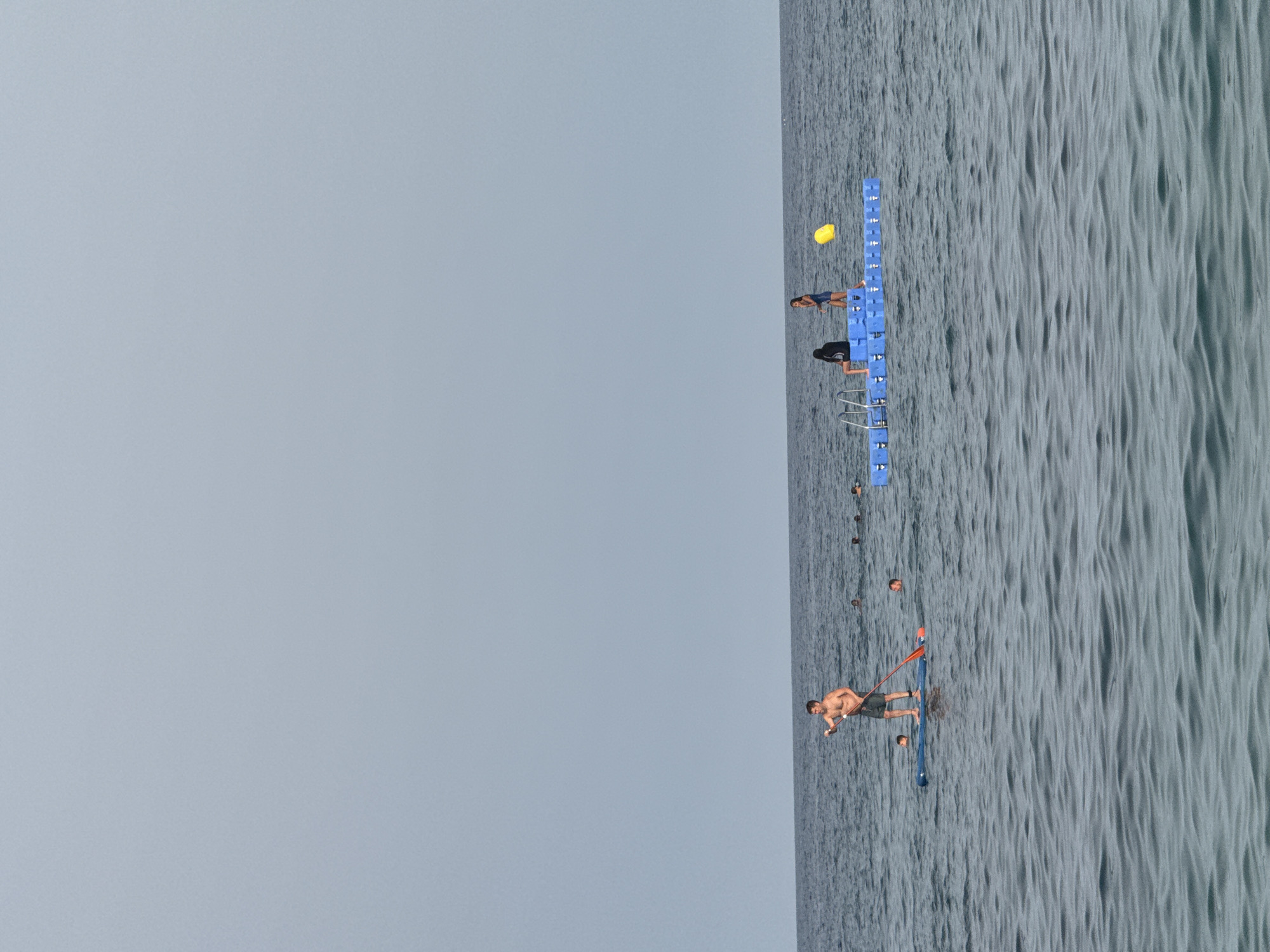 In the waters off the coast of Platja d'Aro, Spain, a group of people are enjoying recreational activities. A man stands on a paddleboard, holding a paddle, in the foreground. In the middle ground, a blue floating platform is visible, with two individuals standing on it and one person sitting. Several other people are swimming in the water around the platform and paddleboarder. A yellow buoy is also present in the water near the platform. The sea is calm, with small waves, and the sky is overcast. The overall scene depicts a typical summer day at the beach with various water activities.
