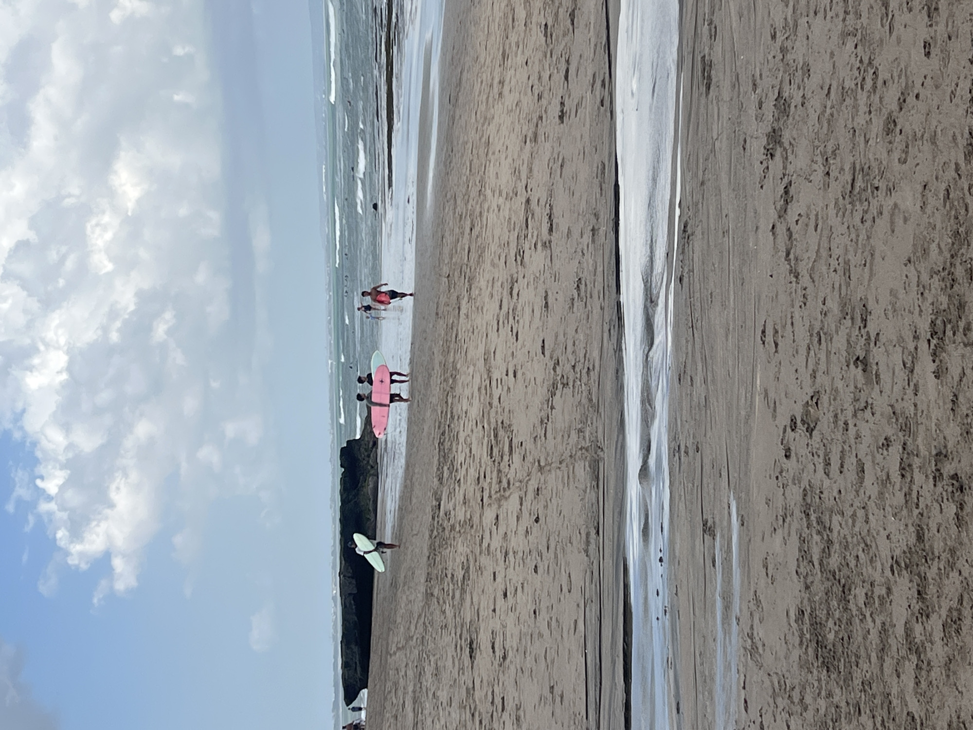 The image shows a beach scene at Kuta, North Kuta, Indonesia. In the foreground, a wet sand area with shallow water pools and sand tracks is visible. The midground is a sandy beach where several individuals are walking. Two people are carrying surfboards, one pink and one light blue. Further back, another person is walking with a surfboard. In the distance, there are more people in the ocean, some on surfboards, and waves are breaking. The background features a rocky outcrop on the left and the ocean with a cloudy sky above.