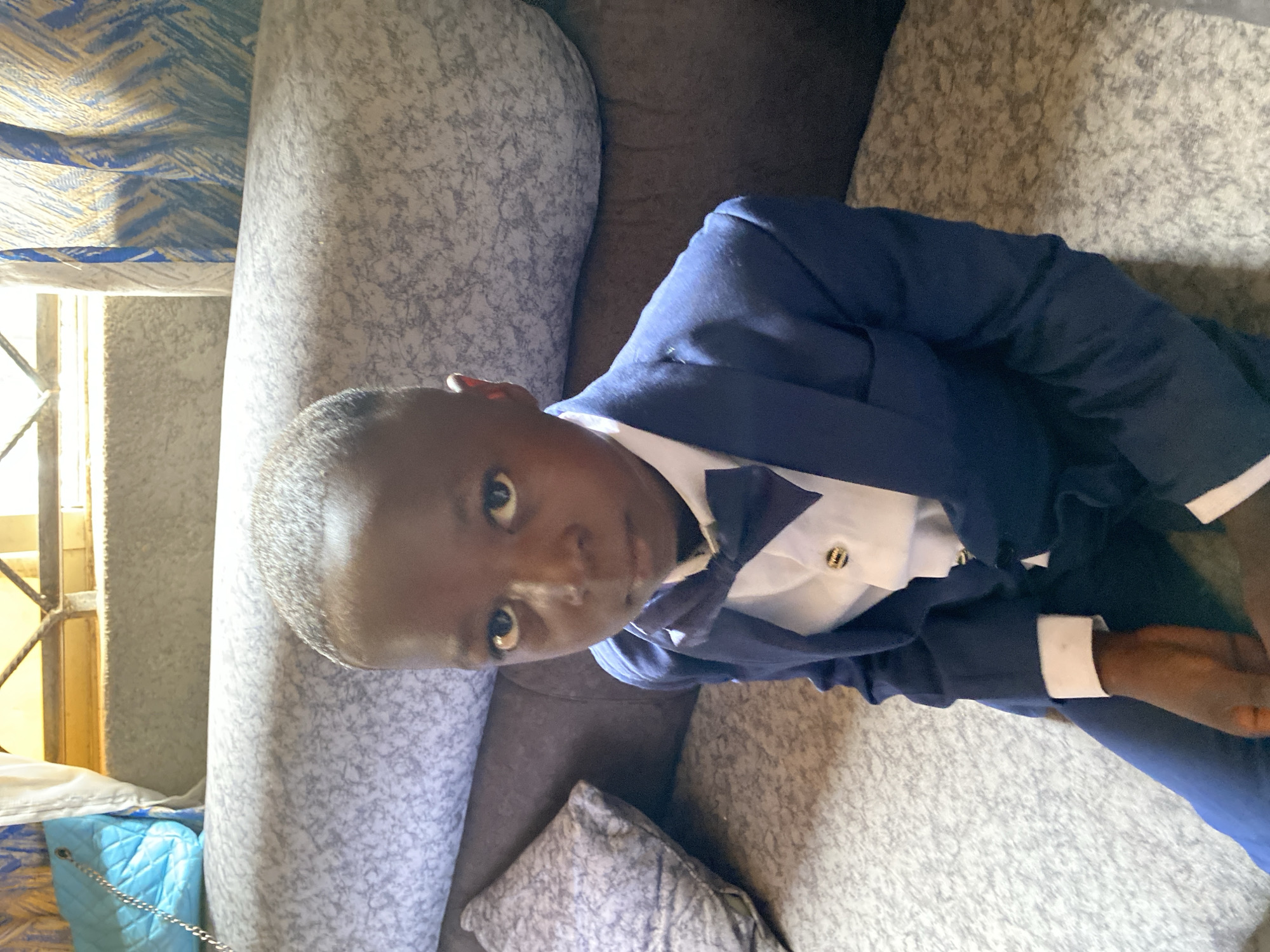 A young boy is lying on a light gray sofa with a dark brown cushion, wearing a navy blue suit with a white shirt and a bow tie. The scene appears to be indoors, near a window with patterned curtains. The image appears to be taken inside an apartment. Kuru Dungus, Nigeria, is not explicitly apparent in the image.