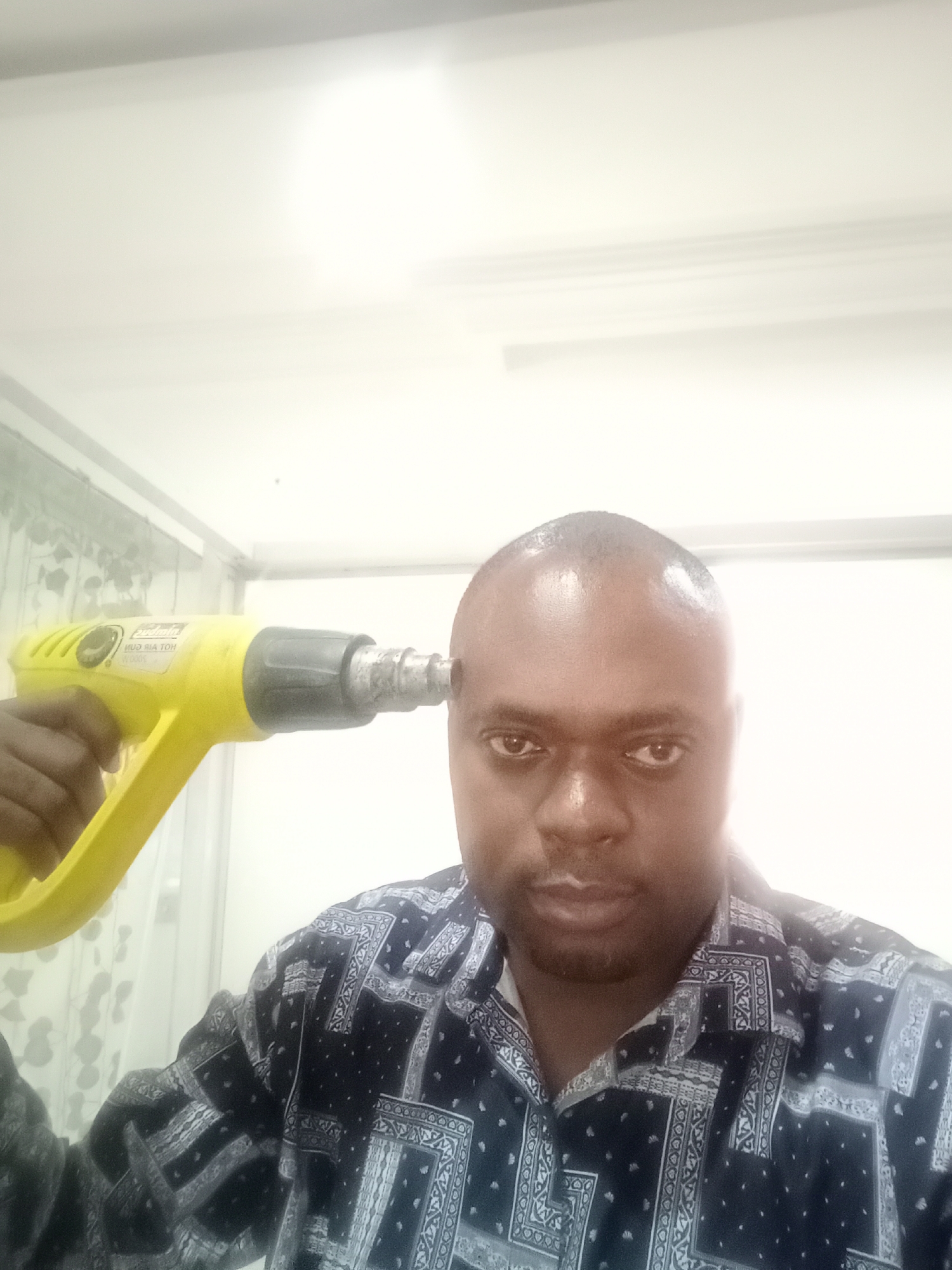 The image depicts a waist-up portrait of a man in an indoor setting, likely in Gwarinpa, Nigeria. The man has a dark complexion and a bald head, looking directly at the camera with a serious or neutral expression. He is wearing a dark blue or black shirt adorned with a white and grey patterned design, possibly geometric or paisley.
In his right hand, the man holds a bright yellow hot air gun (heat gun), the nozzle of which is pressed against his right temple. The visible text on the side of the tool clearly reads "HOT AIR GUN" and "2000W," indicating its function and power. A brand name, possibly "Zulala," is also faintly discernible. This pose suggests a staged or symbolic act, humorously or dramatically mimicking pointing a weapon at oneself, rather than an actual act of harm.
The background is predominantly bright white, suggesting strong lighting or an overexposed setting, possibly a white wall or ceiling. A faint, light-colored patterned curtain or wallpaper is visible on the left side of the frame, behind the man's left shoulder and the heat gun. The bright lighting conditions make it difficult to ascertain the time of day, though it appears to be well-lit artificially. The focus is sharp on the man and the tool, making them the central subjects of the image.