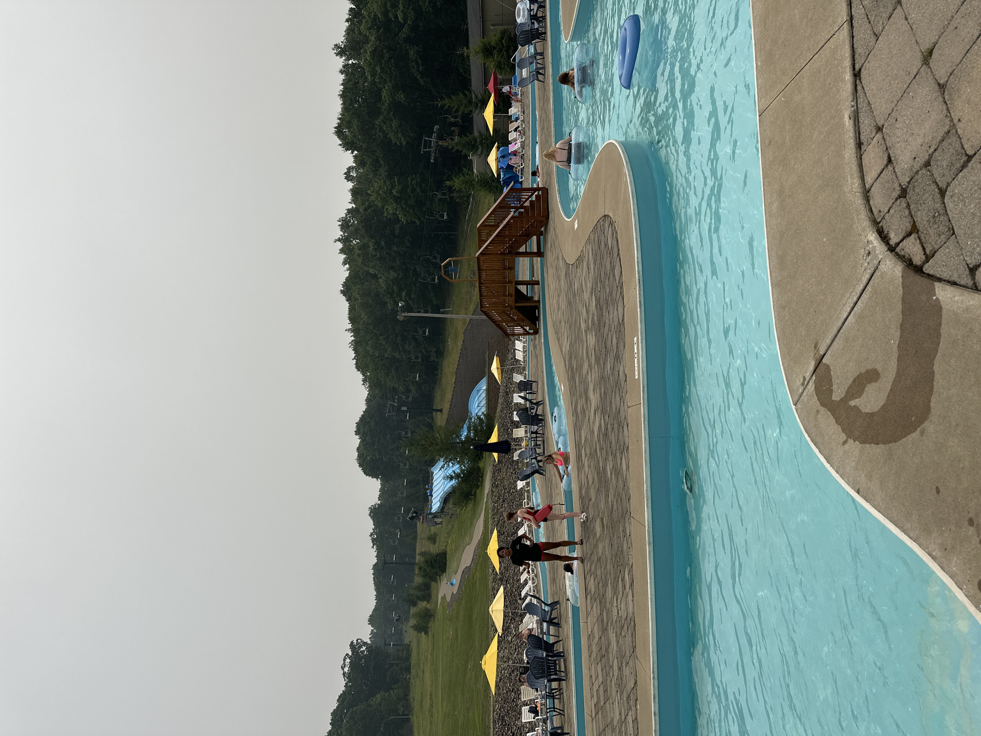 The image depicts a water park on a hazy day, with a lazy river filled with people relaxing in inner tubes. The lazy river has bright blue water and is bordered by concrete and brick pathways. In the background, a blue water slide is visible on a grassy hill, and ski lifts are present on the higher slopes, suggesting the location may also function as a ski resort during colder months. Several lounge chairs and umbrellas are set up around the pool area. Two individuals, possibly lifeguards, are standing near the edge of the river, one in a black shirt and shorts, the other in a red swimsuit. Further in the background, a wooden walkway with stairs leads up to an elevated platform. The overall environment appears to be a recreational facility set amidst a heavily wooded area.