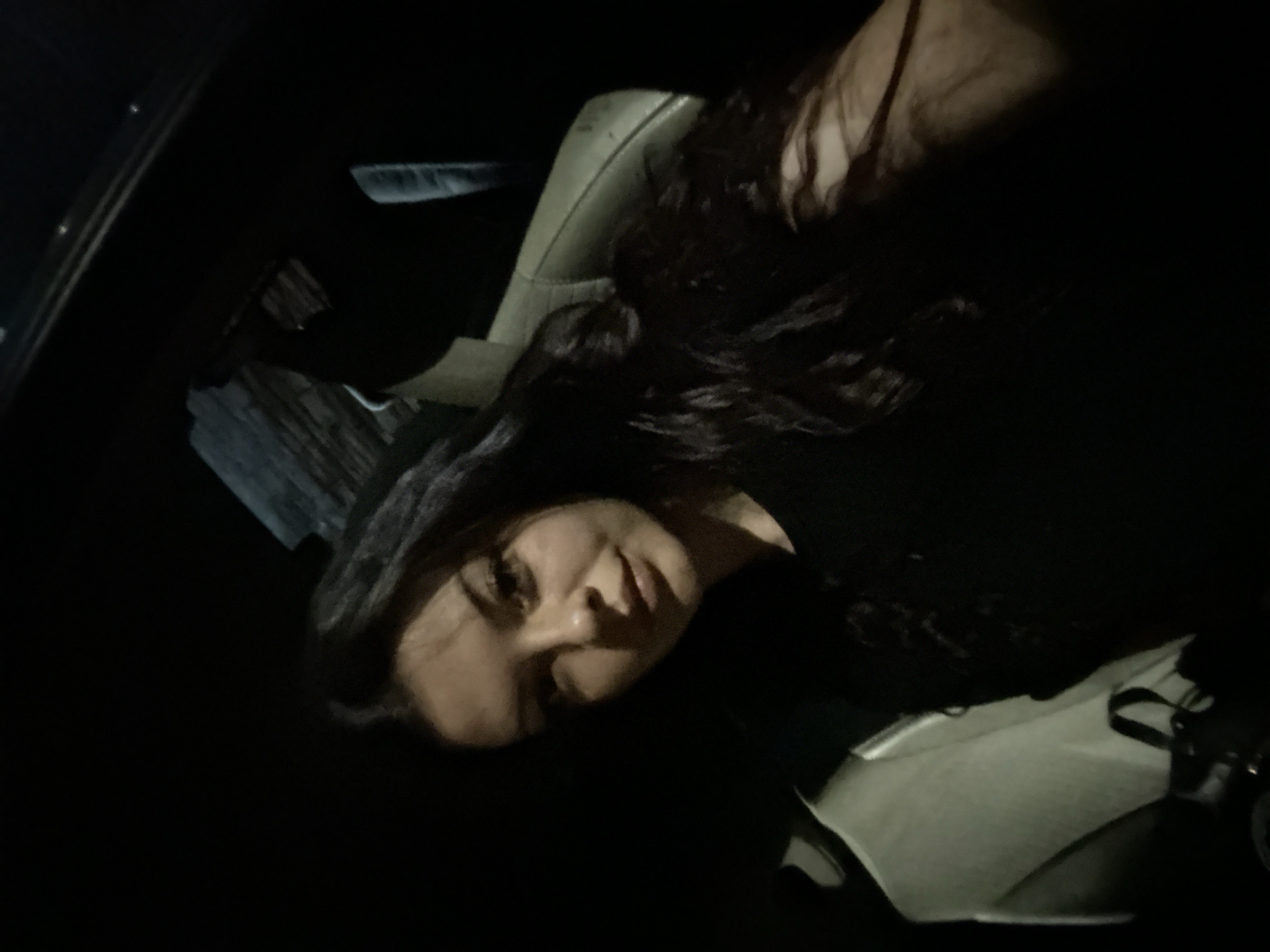 A young woman with long, dark, wavy hair is captured in a selfie taken from a low angle. She is wearing a black top. Her face is partially illuminated, with her eyes looking towards the camera, and a slight smile. The background is dark, suggesting an indoor or nighttime setting, possibly a car interior, indicated by a textured surface that could be a car seat or dashboard visible to her right. Her left side is lighter, with a creamy-colored object that might be another person's arm or a bag. The image's quality is grainy and low-light, characteristic of a photograph taken in dim conditions, potentially in Sliven, Bulgaria.