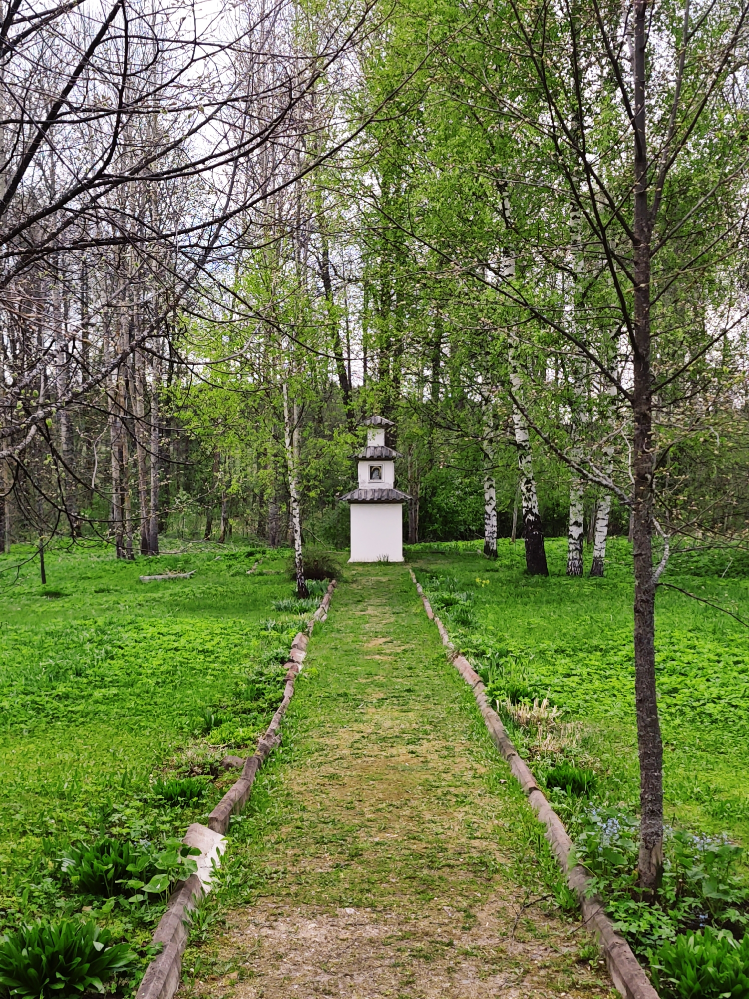 The image depicts a path leading to a small, white, multi-tiered structure in a green, grassy area with trees, possibly a park. The location is identified as Spas-Ugol, Russia. The path has a border of concrete blocks or low walls along its sides. Trees with green foliage are present on the right, while the left side has more bare trees or trees with less developed foliage. The sky is not visible. No people or animals are observed.