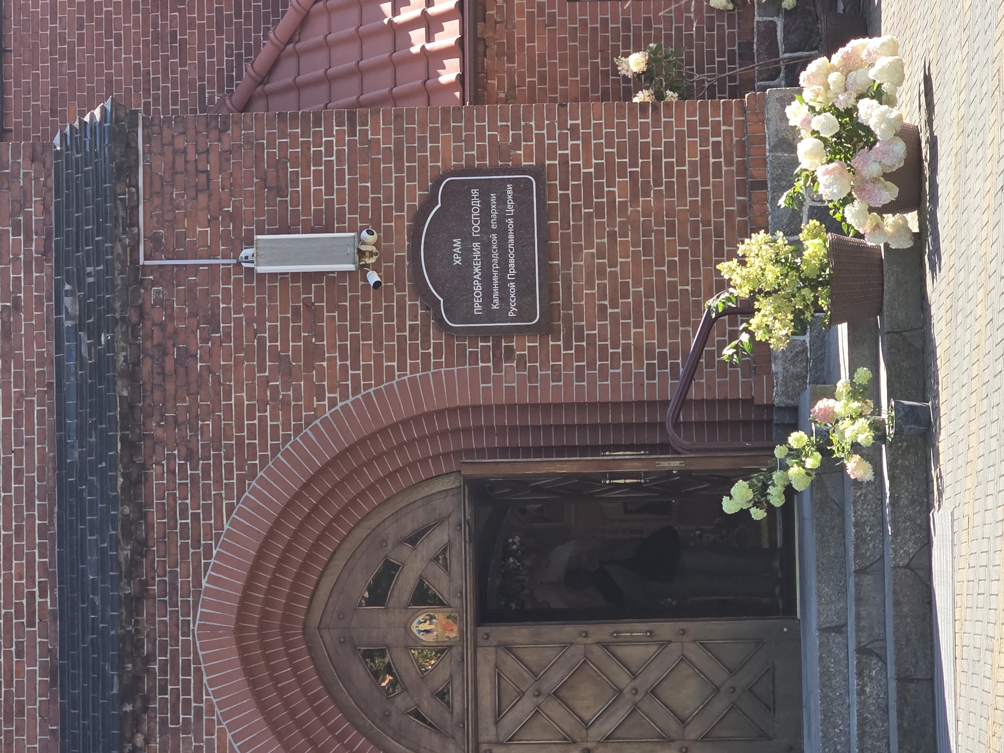The image shows the entrance of a brick church in Zelenogradsk, Russia. The church has a large, ornate wooden door with a semicircular archway. A sign above the door reads "ХРАМ ПРЕОБРАЖЕНИЯ ГОСПОДНЯ Калининградской епархии Русской Православной Церкви" which translates to "Church of the Transfiguration of the Lord of the Kaliningrad Diocese of the Russian Orthodox Church". Two security cameras are mounted on the wall above the sign.
Steps lead up to the entrance, decorated with potted hydrangeas. The exterior walls of the church are made of red brick, and the roof is covered with terracotta tiles. The surrounding area is paved with light-colored bricks, and a metal railing runs along the side of the steps. The sky is clear and bright, indicating a sunny day.