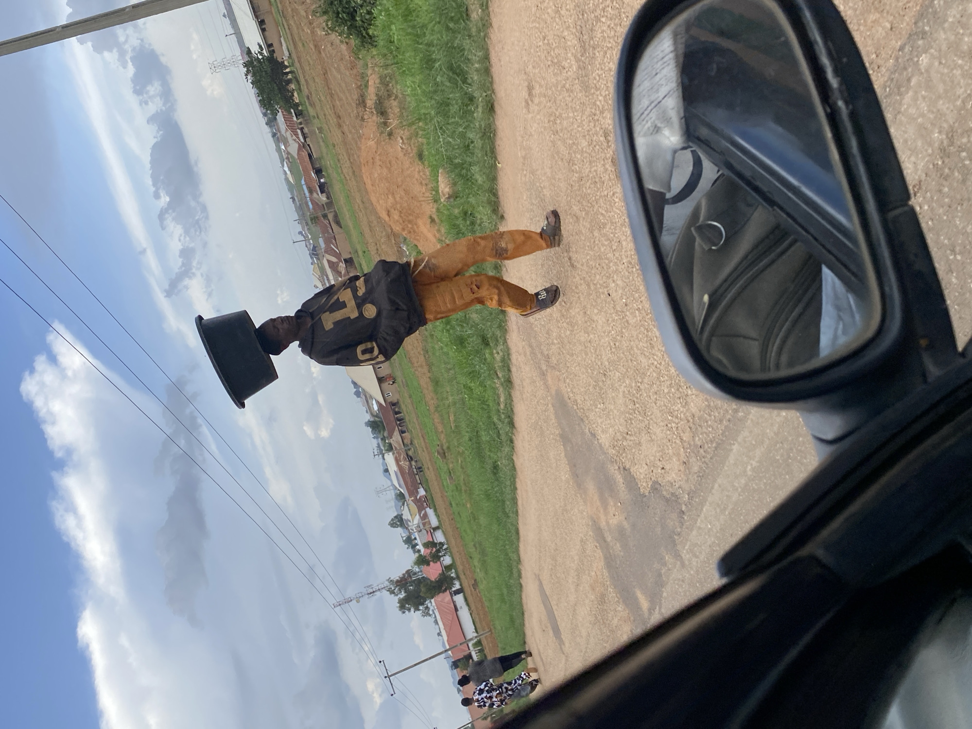 The image shows a young Black man walking on a dirt road, carrying a black tub on his head. He is wearing a black hooded sweatshirt with a gold logo and orange pants. In the background, there are buildings, trees, and a cloudy sky. There are also power lines and a cell tower visible. Two other people are walking in the distance on the left side of the road. A car's side mirror is in the foreground on the right, reflecting the interior of the car. The scene appears to be in a rural or semi-urban area. Based on the visual cues of the architecture, vegetation, and the appearance of the people, the location is identified as Ban, Nigeria.