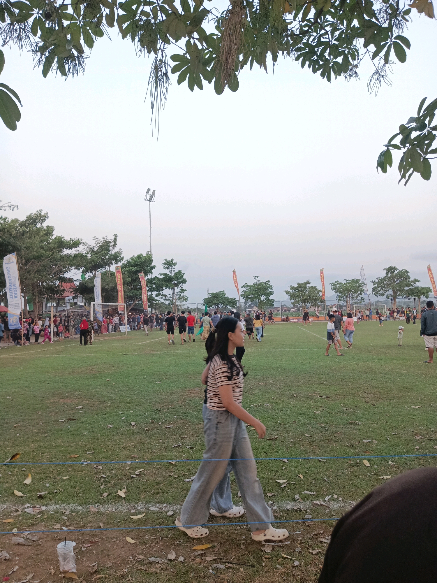 The image depicts an outdoor event, likely a community gathering or sporting event, at a grassy field in Tawing, Indonesia. In the foreground, two individuals are walking across the field, one of whom is a young woman wearing a striped t-shirt and wide-leg jeans. Her companion's back is to the camera. In the background, a crowd of people are gathered, some near a soccer goal and others dispersed across the field. Several flags with text are visible, suggesting sponsorship or organizational presence. Trees line the perimeter of the field, and a light pole with floodlights stands tall in the distance. The sky is overcast, indicating potentially late afternoon or early evening. The overall impression is one of a casual, public event taking place in a semi-urban or suburban setting.