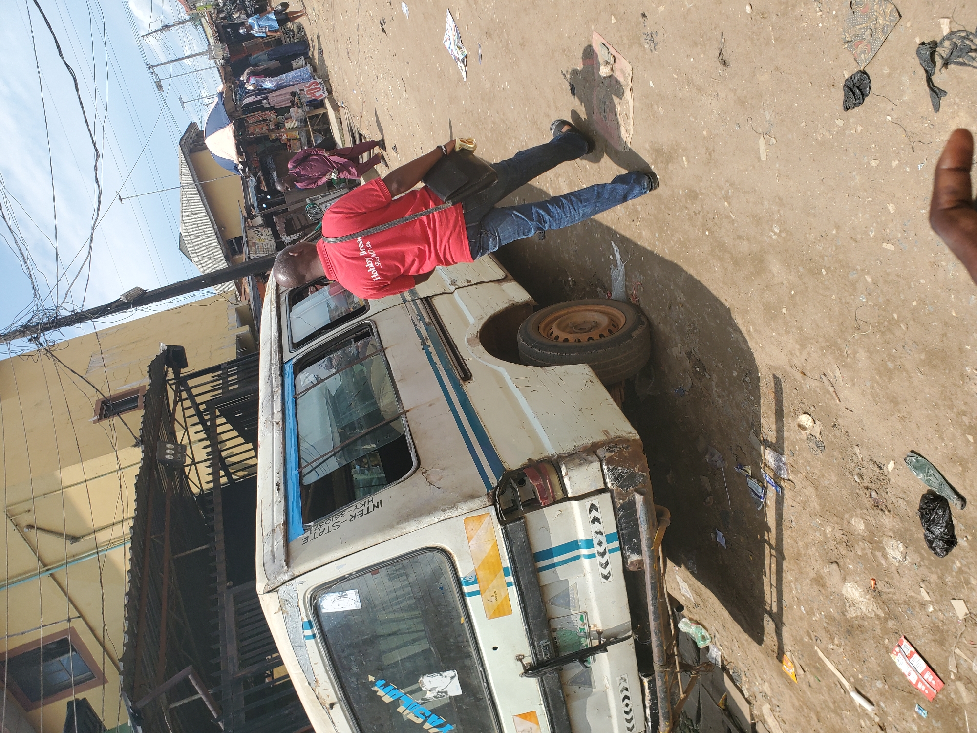 The image depicts a scene in the city of Shangisha, Nigeria. A white van, appearing worn and dirty, is parked on a dusty, unpaved road. The van has blue stripes along its side and a license plate that is partially visible. A man wearing a red polo shirt with "Hobby Brea" visible on it and blue jeans is walking away from the van, carrying a black bag over his shoulder. He is wearing sandals. In the background, there are buildings, market stalls with clothing displayed, and other people. The environment appears to be a bustling urban area with overhead wires and a bright, sunny sky. The ground is littered with debris, including plastic bags and papers.