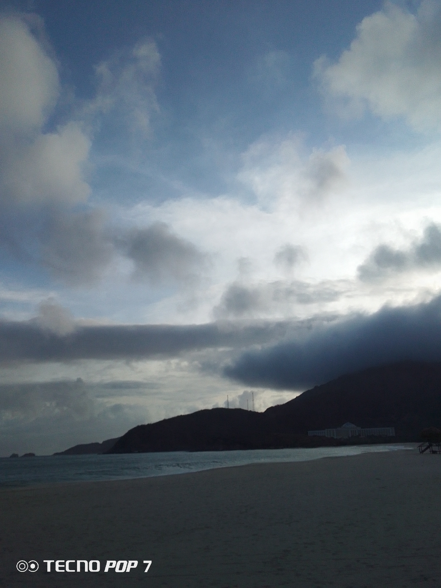 The image presents a serene coastal landscape in Pedro González, Venezuela, captured under what appears to be an overcast sky or during dawn/dusk. The foreground is dominated by a wide, pale sandy beach that meets the calm, blue-grey waters of the ocean.
In the middle ground, dark, undulating mountains rise sharply from the coastline. Heavy, dark grey clouds partially obscure their peaks and lower slopes, casting shadows over the terrain. A large, light-colored building, likely a resort or hotel, is visible at the base of one of the mountains, standing out against the darker surroundings. To the far left, small rocky islets dot the sea, while to the far right on the beach, an indistinct dark structure, possibly a small shelter, can be faintly seen.
The sky is a dramatic mix of colors, with areas of bright white and light blue peeking through thick, dark grey cloud formations. The overall lighting suggests a subdued atmosphere, lending a tranquil yet grand feel to the scene, with a hint of drama from the heavy cloud cover. No people or specific activities are visible. A watermark "OO TECNO POP 7" is present in the bottom left corner of the image.