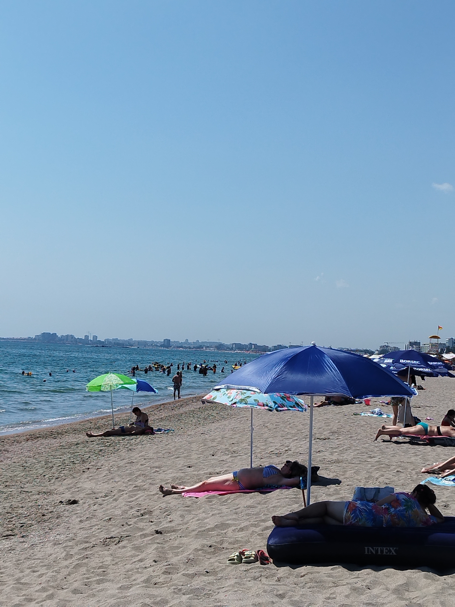 A vertical, eye-level, medium shot captures a lively beach scene on a sunny day, with the city of Constanța, Romania, visible in the background. The foreground is dominated by a sandy beach where several people are relaxing and sunbathing. Two women are the most prominent figures in the foreground, each lying on a towel or air mattress. The woman on the left, in the mid-ground, is lying on her back on a pink towel, wearing a blue and white striped bikini. To her right, in the foreground, another woman is lying on an inflatable air mattress with "INTEX" printed on it, wearing a colorful, floral-patterned top and patterned shorts. She is propped up on her arm, looking to her right.
Further back, a large blue umbrella provides shade for a patterned beach blanket, under which another person is partially visible. To the left of the blue umbrella, a green and blue umbrella casts shadows on the sand, with a person sitting up on a towel beneath it. The sand is a light brown and appears dry.
In the mid-ground, numerous people are seen enjoying the water, swimming and wading in the wavy, blue sea. The shoreline is marked by gentle waves lapping onto the sand. In the distance, across the water, the skyline of Constanța is visible, featuring a collection of buildings and structures. The sky is a clear, bright blue with no clouds, indicating fair weather. The overall impression is one of a busy summer day at the beach.