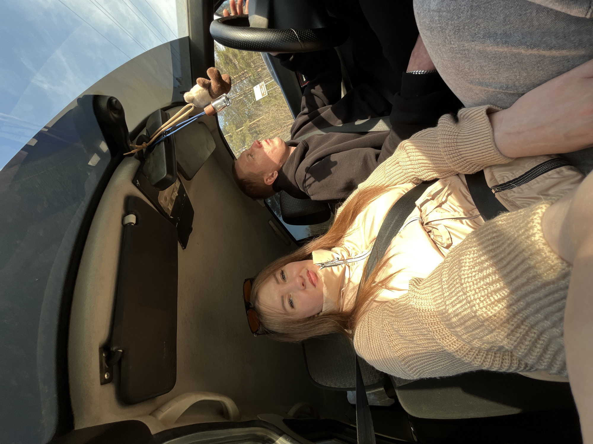 A selfie taken from inside a vehicle, possibly a car, featuring two individuals and part of a third. The first individual, likely the one taking the photo, is a young woman with long blonde hair, wearing a beige jacket and a knitted sweater. She is wearing a seatbelt. Sunglasses are positioned on top of her head.
Behind her, another young male individual is visible, wearing a black hoodie and also wearing a seatbelt. He appears to be looking upwards towards the windshield. The hands of a third individual are seen gripping the steering wheel.
The vehicle interior is visible, including the dashboard, sun visor, and a fuzzy object attached to the visor.
The location is not confirmed as Chaikovsky, Russia. Only interior of car and view through window is visible.