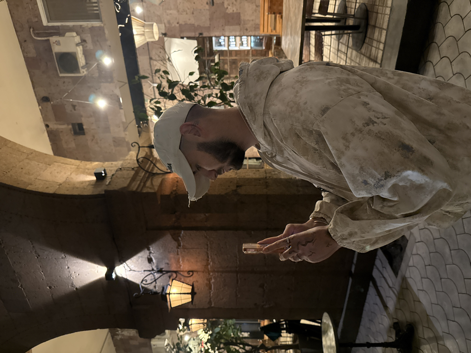 A man with a beard wearing a light-colored baseball cap and a beige and white speckled hooded sweatshirt is holding a small, rectangular object, possibly a phone or wallet, in his hands. He appears to be looking down at the object. His hands have tattoos. He is positioned indoors in what appears to be a restaurant or cafe, judging by the tables and decor in the background. The building's architecture features stone brick walls and arches, along with vintage-style lamp fixtures. Plants are also visible in the background, contributing to the ambiance. The location is Yerevan, Armenia.